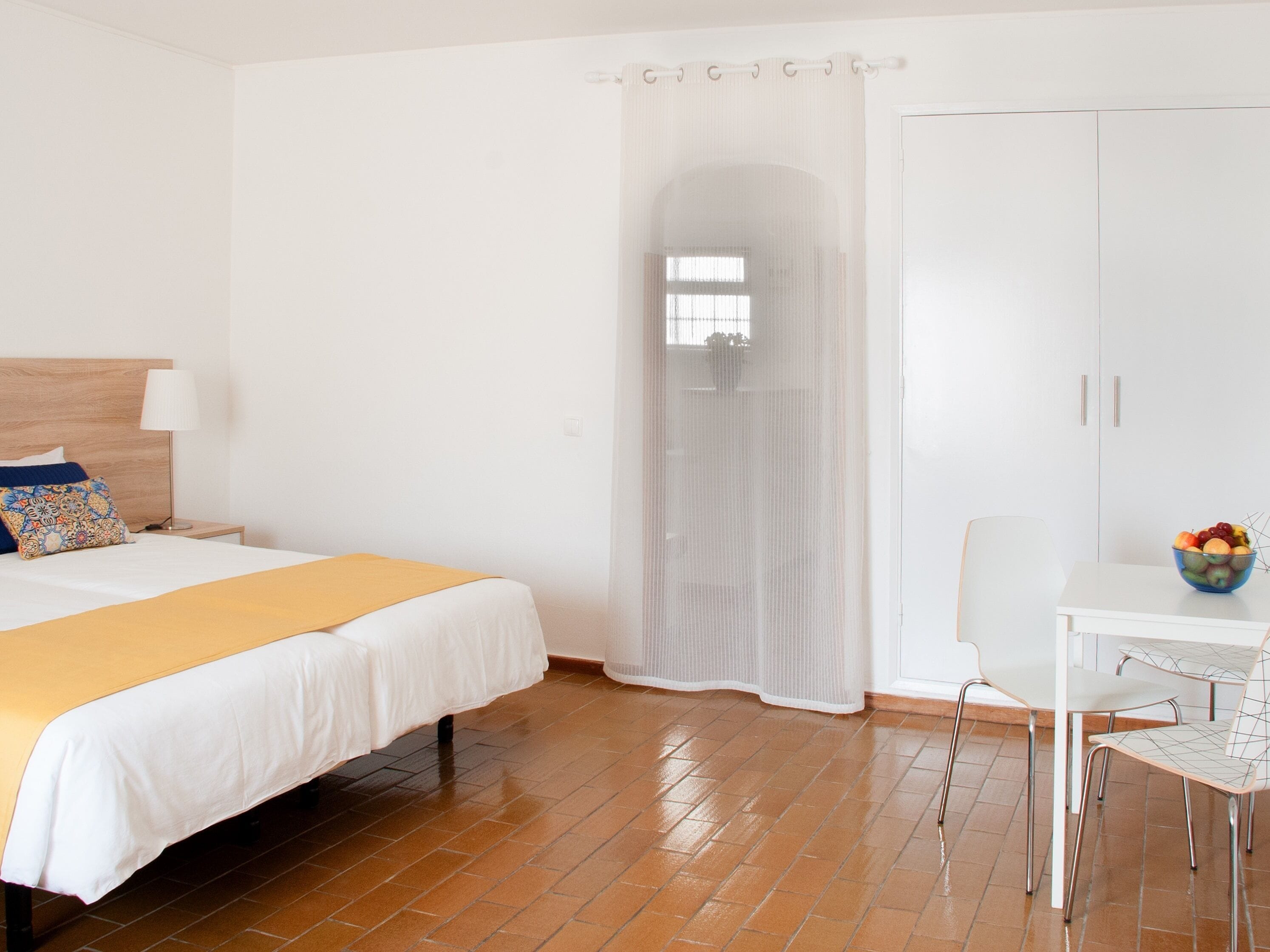 A hotel room with a bed, a nightstand, a table with a bowl of fruit, and a wardrobe.