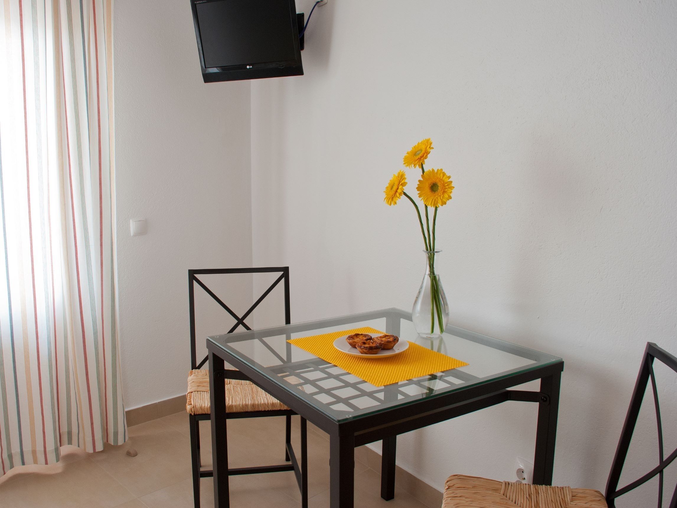 Un'angolare zona pranzo con un tavolo in vetro, due sedie, un vaso con fiori gialli e una televisione fissata alla parete.
