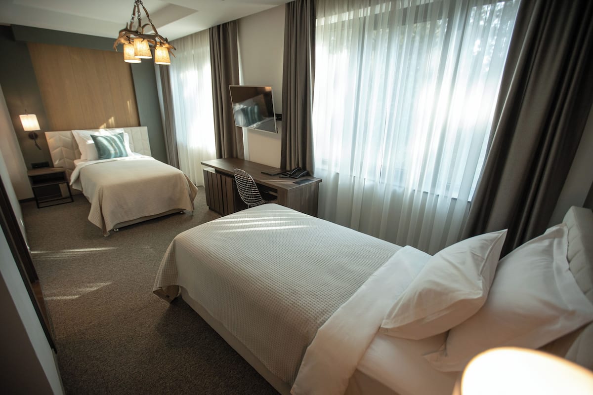 A hotel room with two beds, a desk, a chair, a TV, and a large window with curtains.