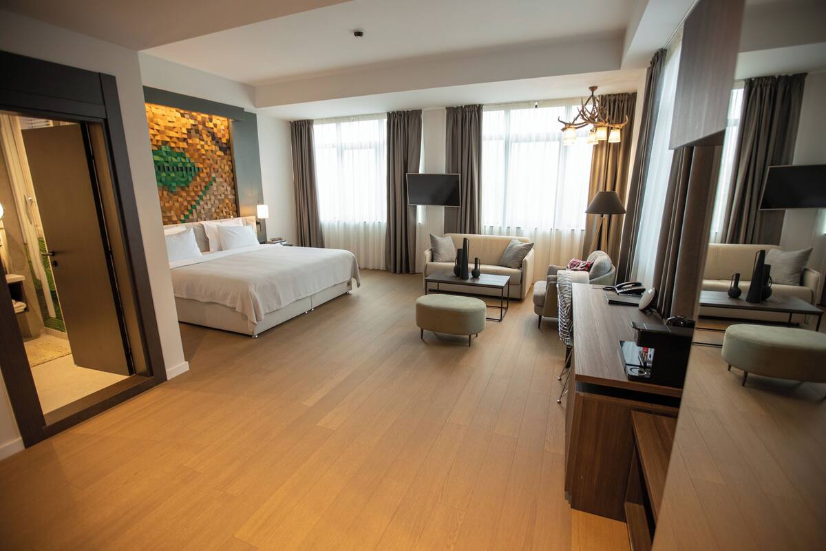A hotel room with a bed, a desk, two armchairs, a sofa, a TV, and a wall art piece.