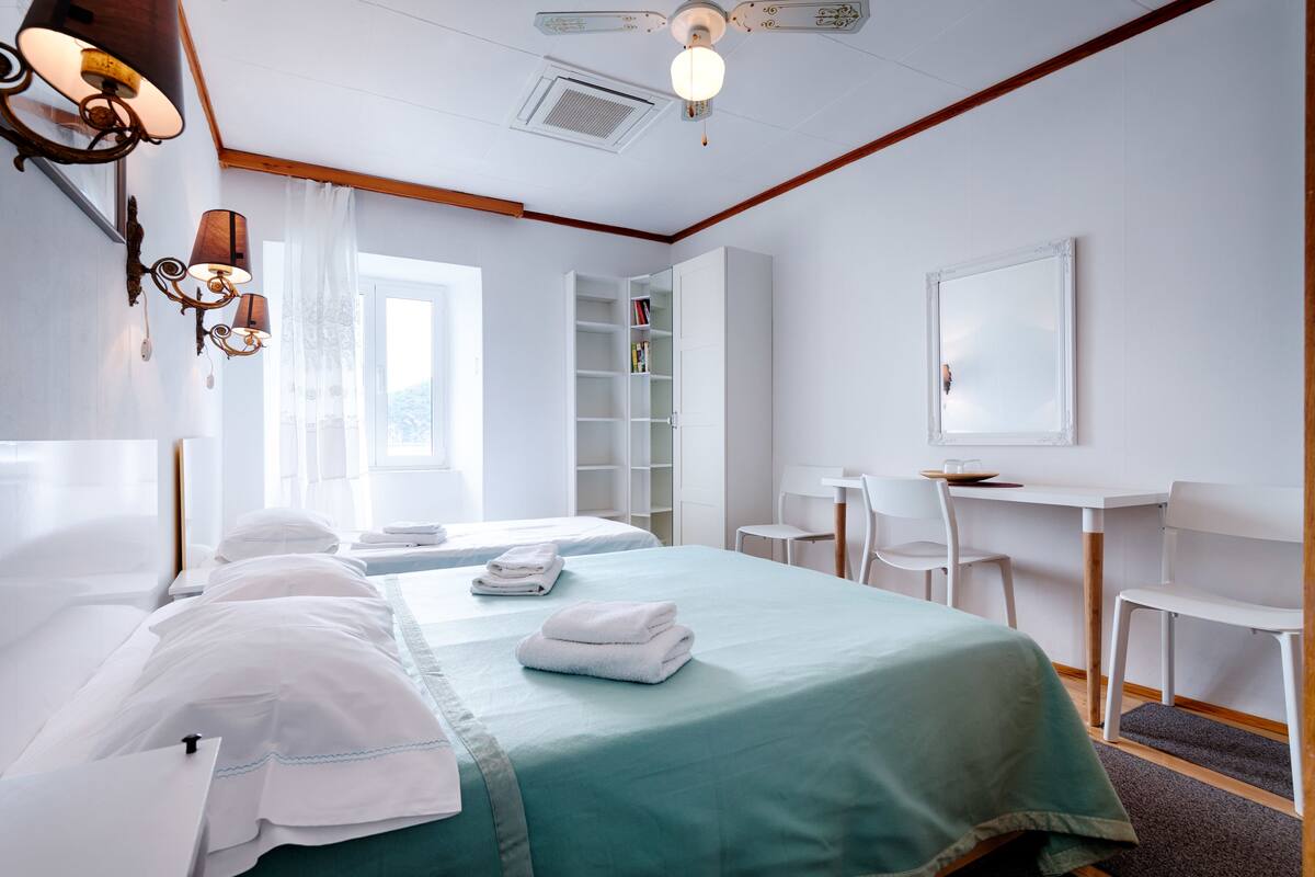 A hotel room with two beds, a desk, a chair, a bookshelf, and a large window.