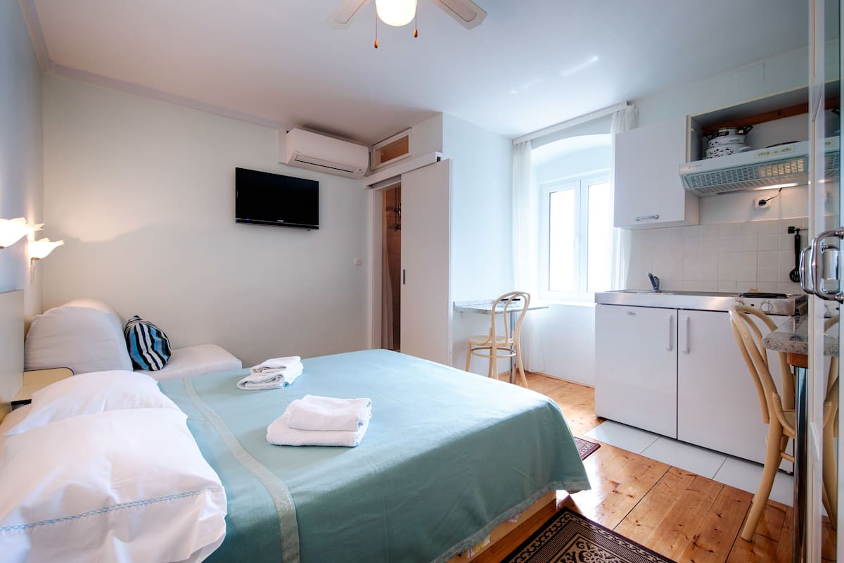 A hotel room with a bed, a desk, a chair, a TV, a kitchenette, and a bathroom.