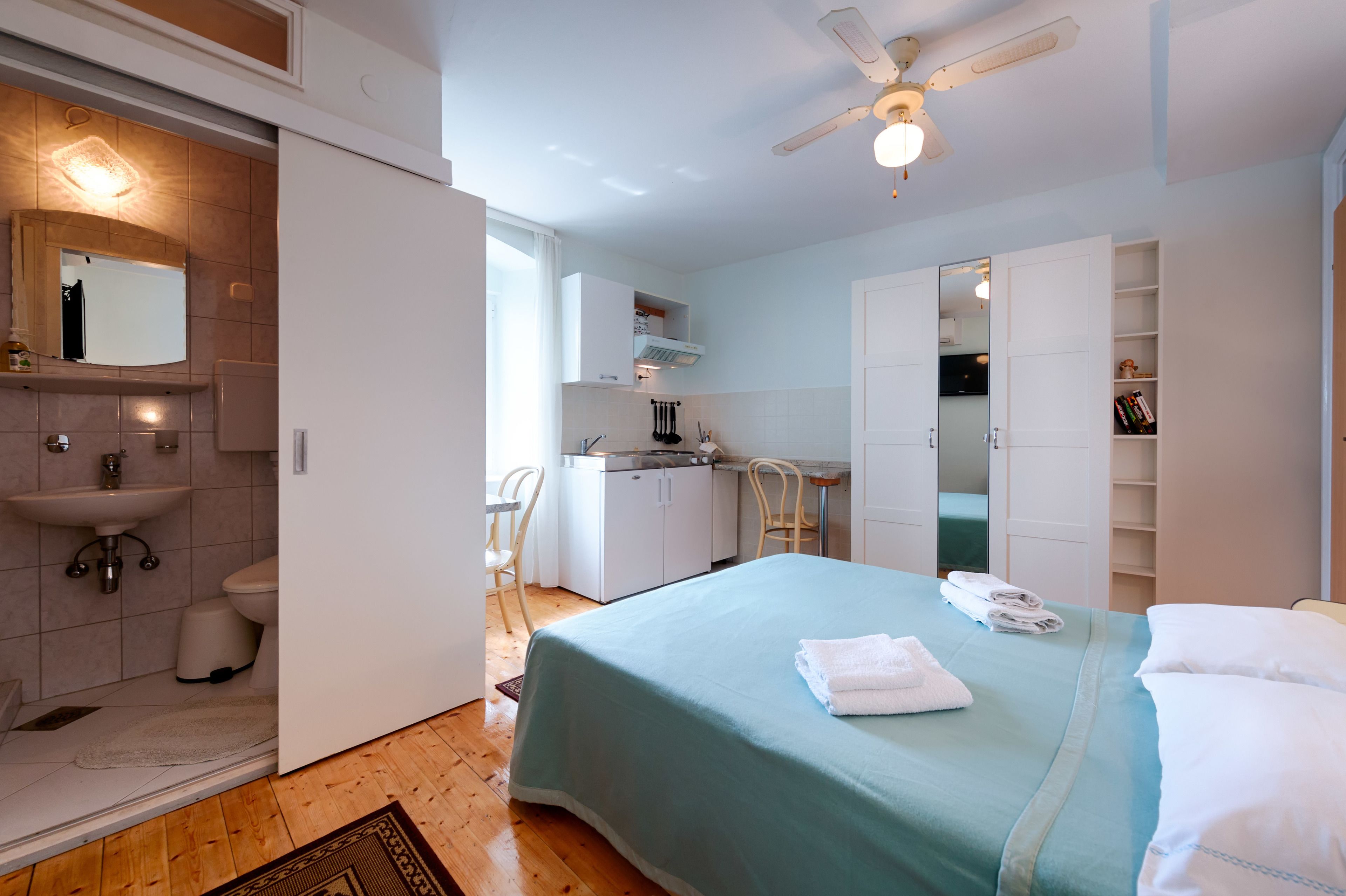 Un dormitorio con cama, ventilador de techo, baño con lavamanos y espejo.