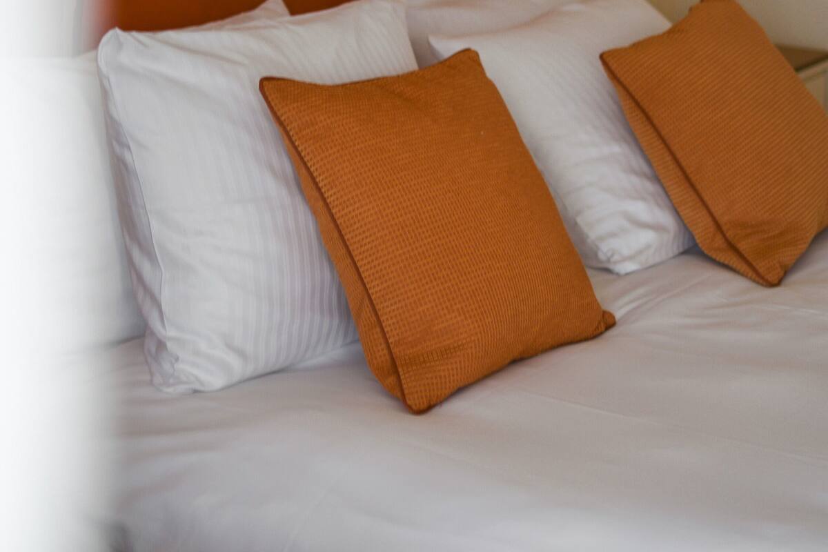 A bed with white bedding and two orange pillows, a wooden headboard, and a wall-mounted lamp.