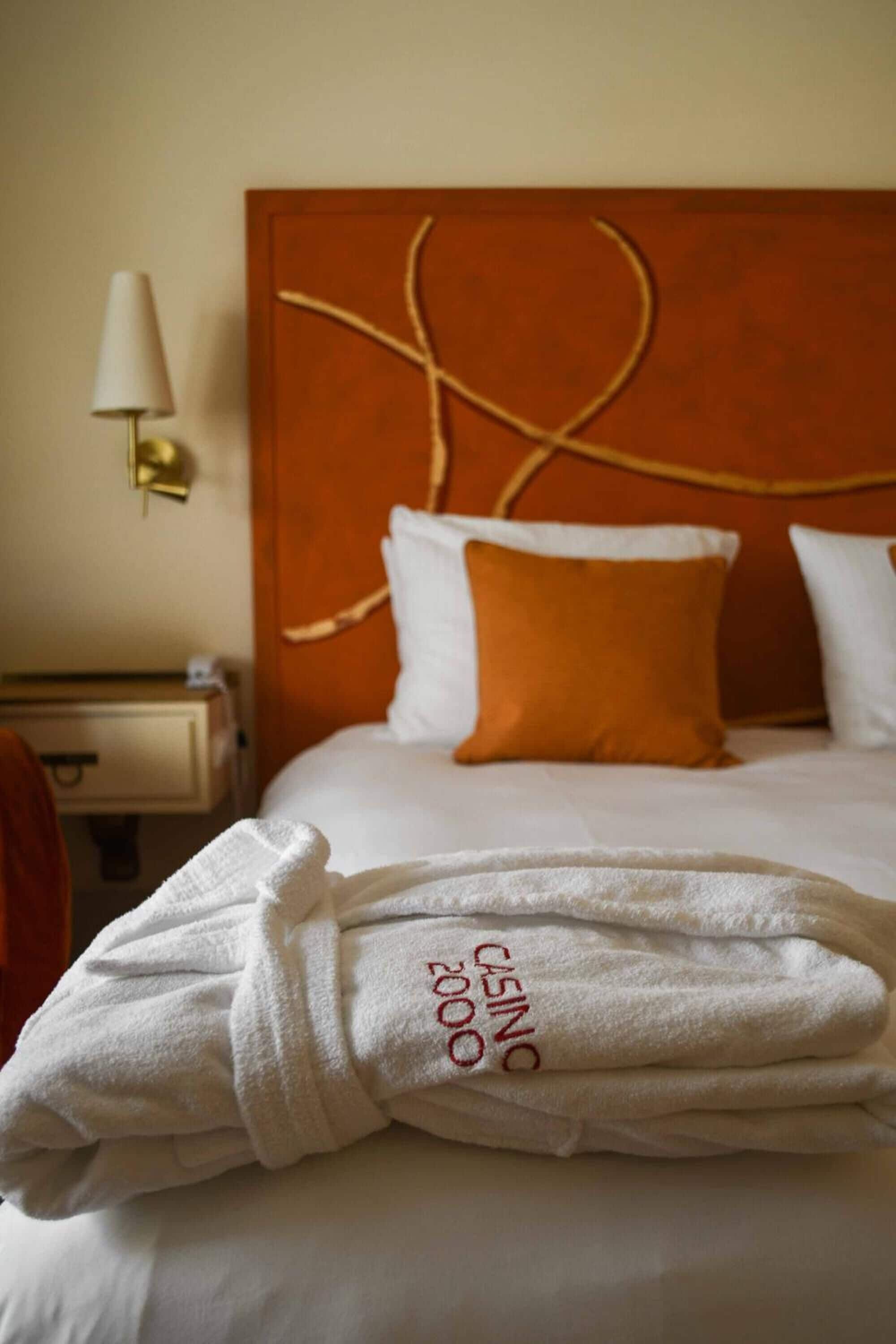 Una cama bien hecha con almohadas blancas y anaranjadas, un albornoz blanco sobre la cama, y una lámpara de pared.