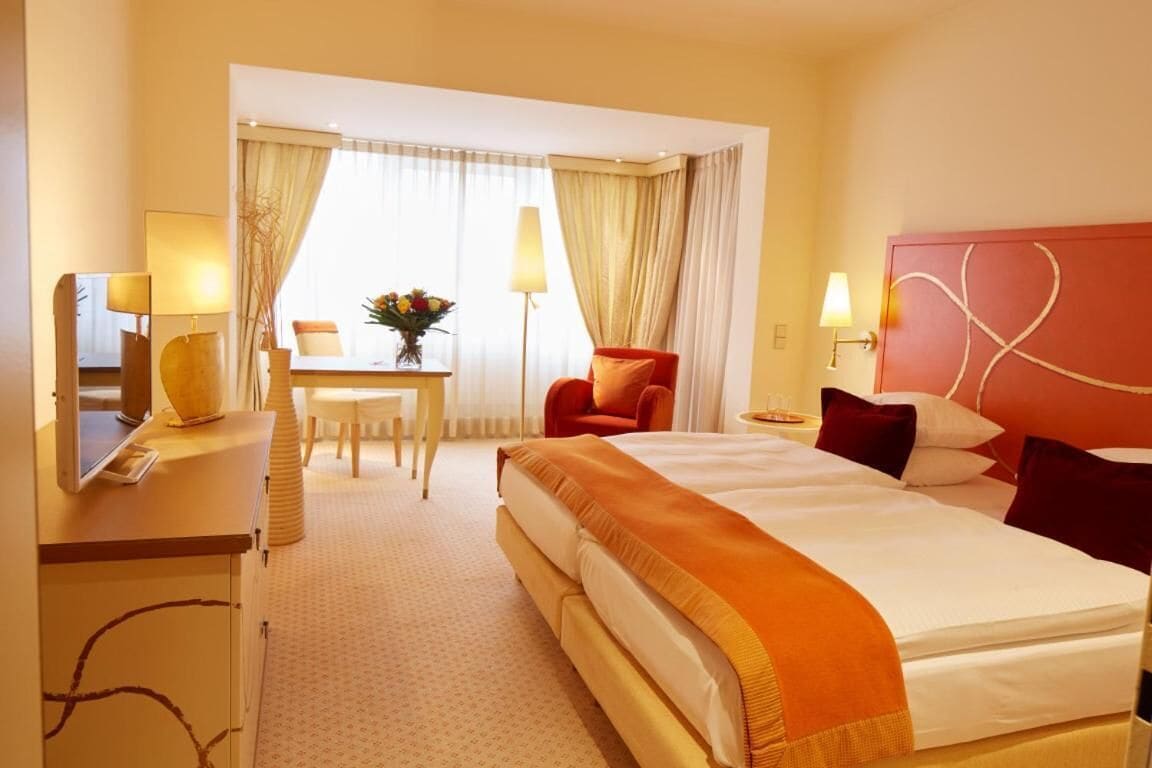 Ein Hotelzimmer mit einem großen Bett, einem Schreibtisch mit Stuhl, einem kleinen Tisch mit Blumenvase und einem roten Sessel.