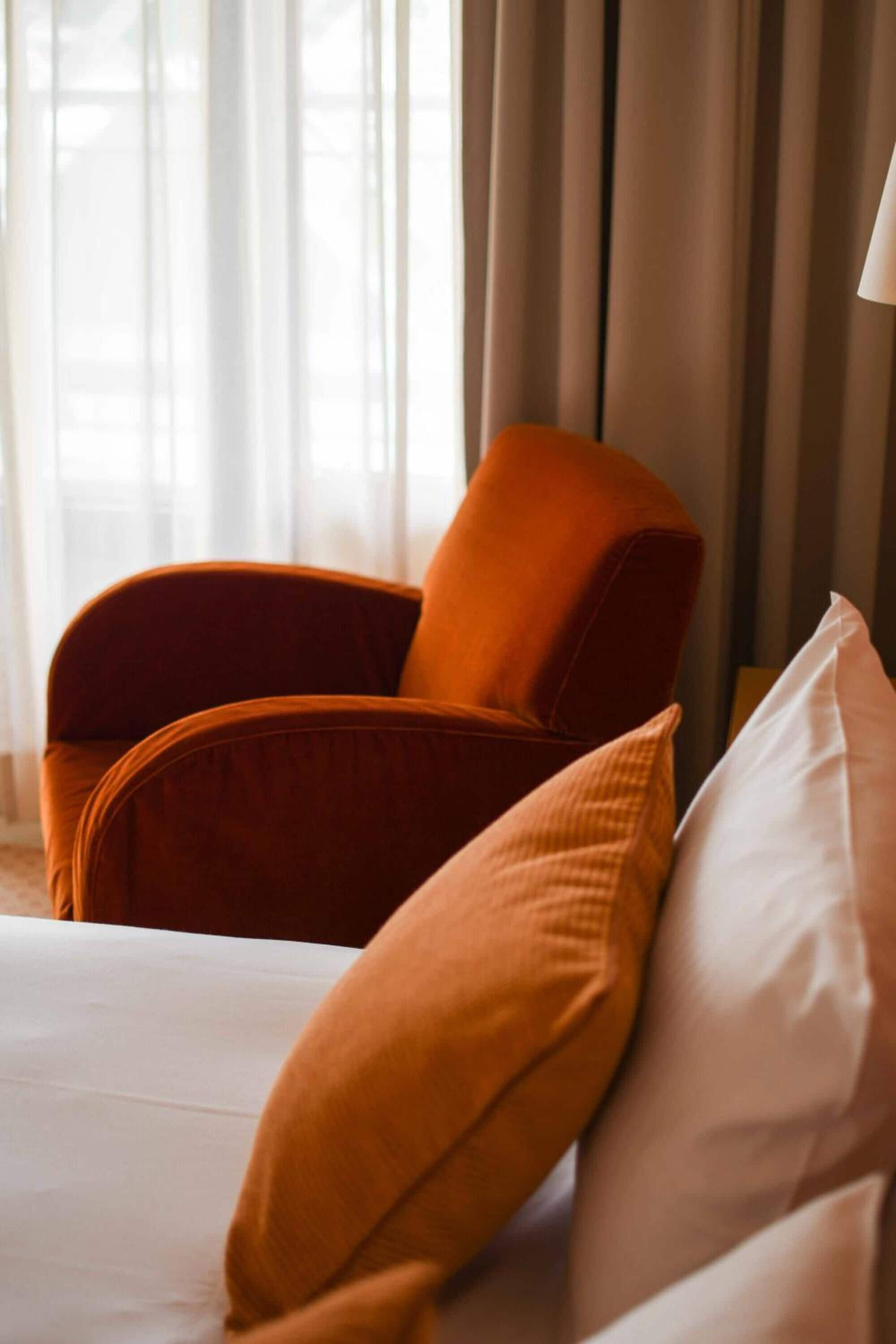 Una habitación de hotel con cama, un ventanal con cortinas y un sillón naranja.