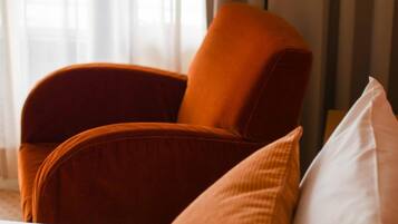 Una habitación de hotel con cama, un ventanal con cortinas y un sillón naranja.