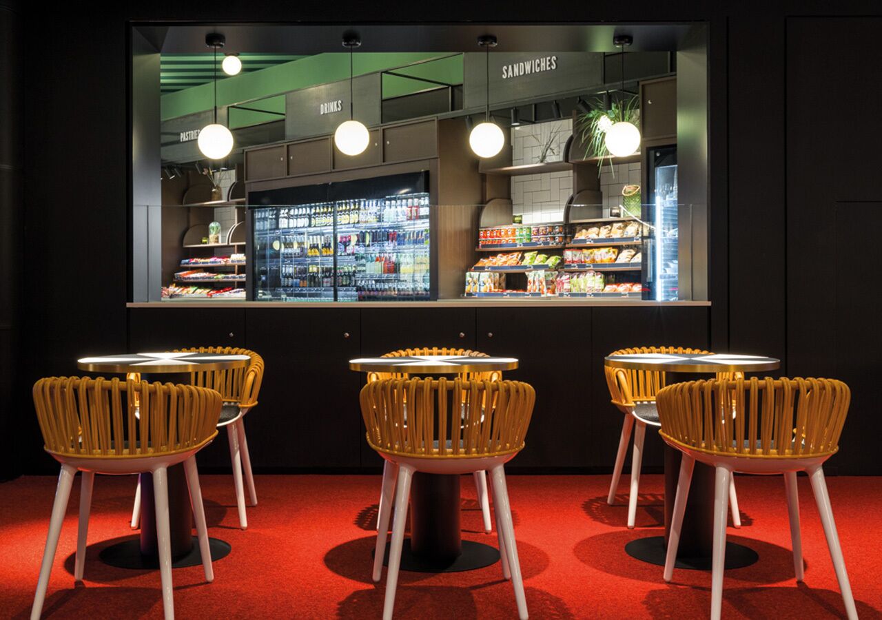 A modern interior with a counter displaying sandwiches and drinks, and three yellow chairs with white legs.