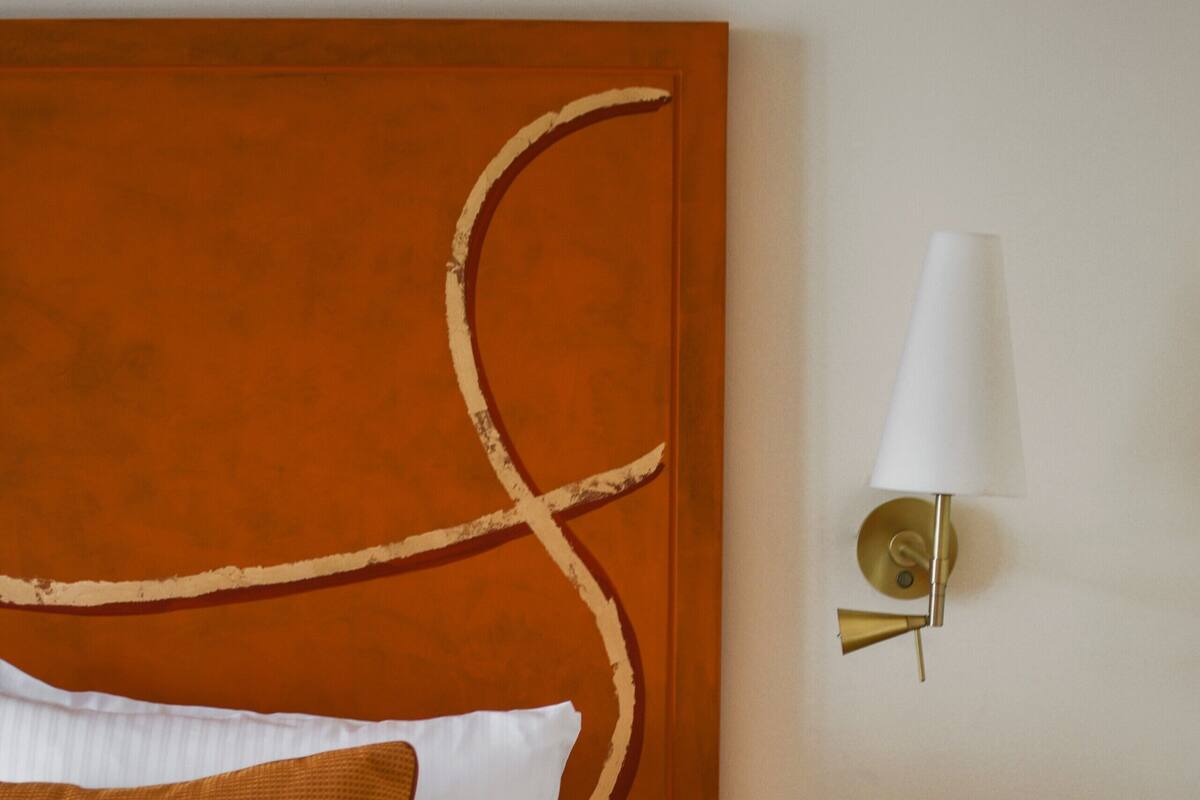 A neatly made bed with white and brown bedding, a wall-mounted lamp, and a small bedside table with a drawer.