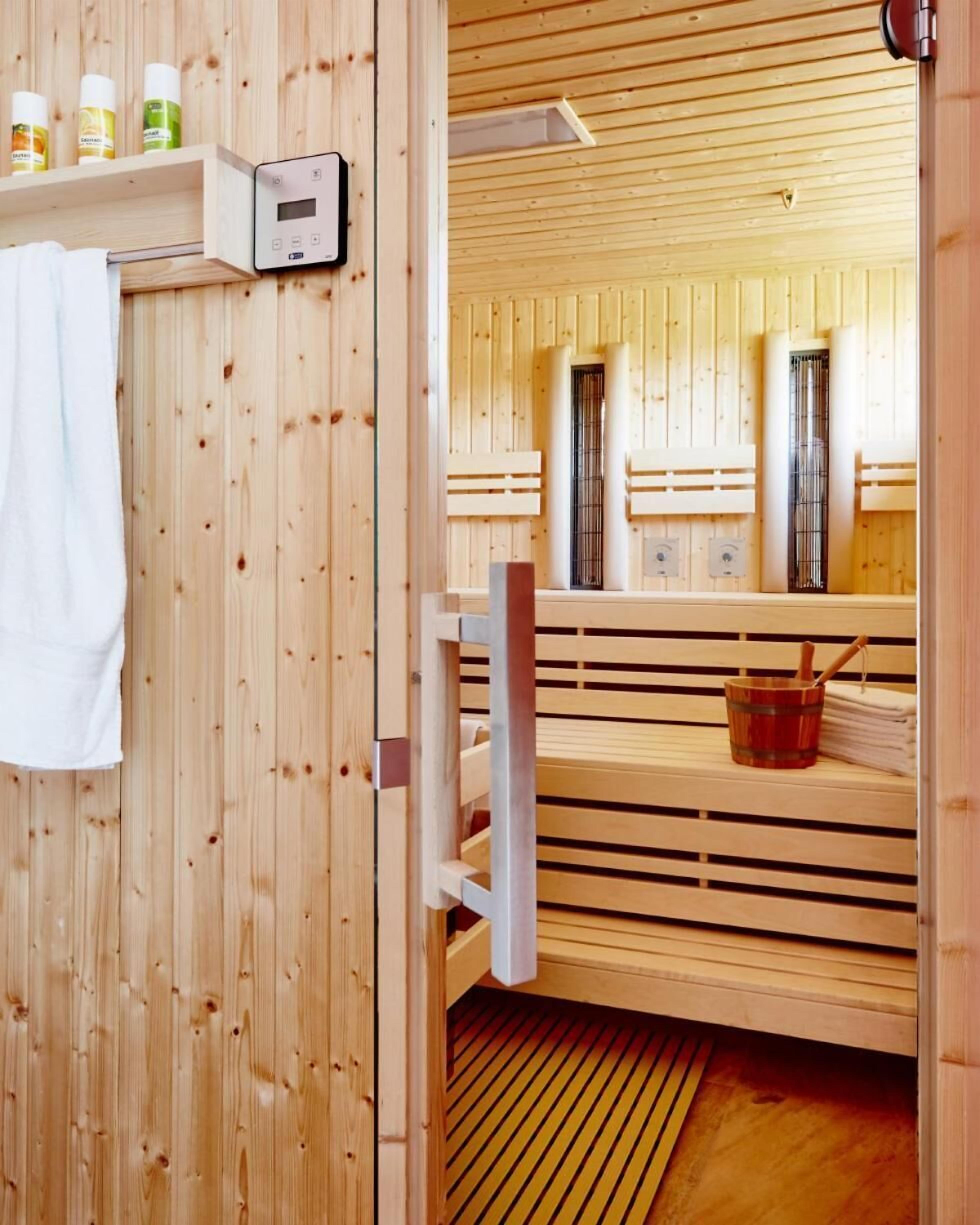 A sauna room with wooden walls and benches. There is a control panel on the wall and a towel hanging on the door.