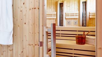 A sauna room with wooden walls and benches. There is a control panel on the wall and a towel hanging on the door.