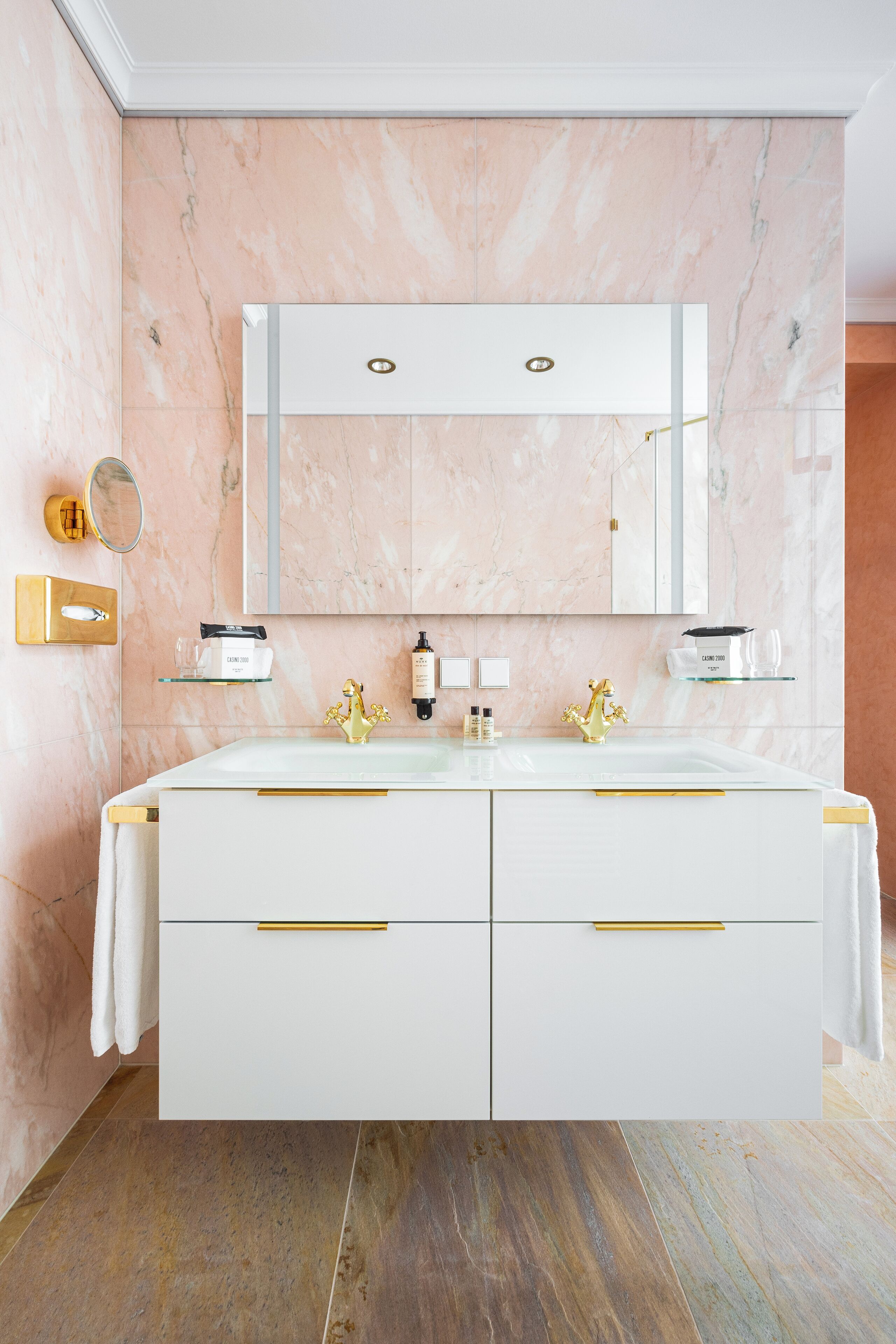 A bathroom with a marble wall, a double sink vanity, gold handles, and a large mirror.