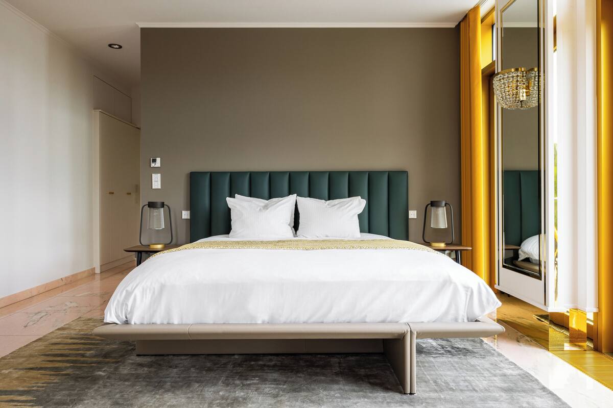 A modern hotel room with a large bed, a green upholstered headboard, and a large mirror with a gold frame.