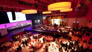 Un gran evento en interiores con varias mesas redondas, un escenario y una pantalla grande que muestra un paisaje urbano.