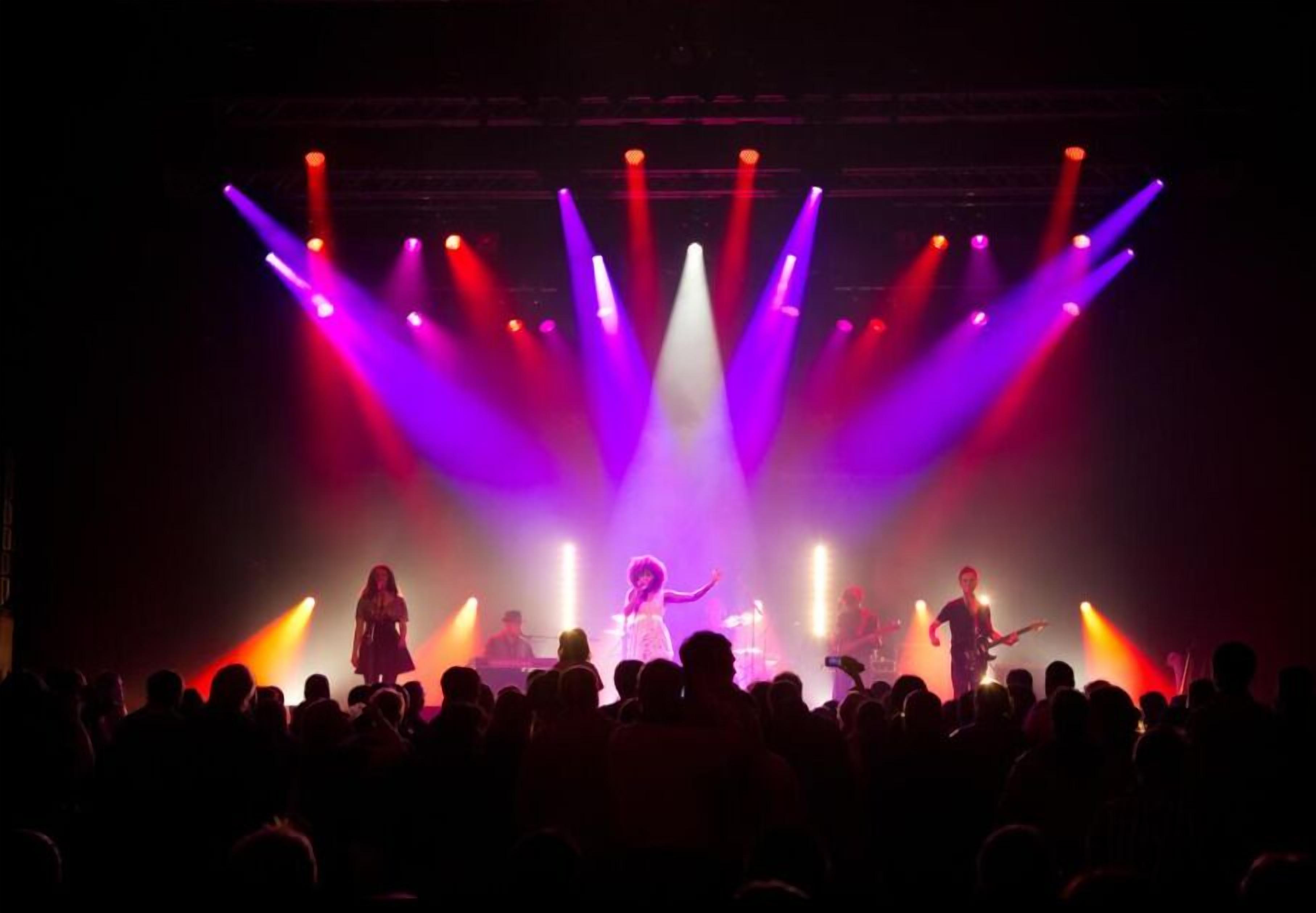 A concert with a band performing on stage, illuminated by colorful lights, and an audience in front.