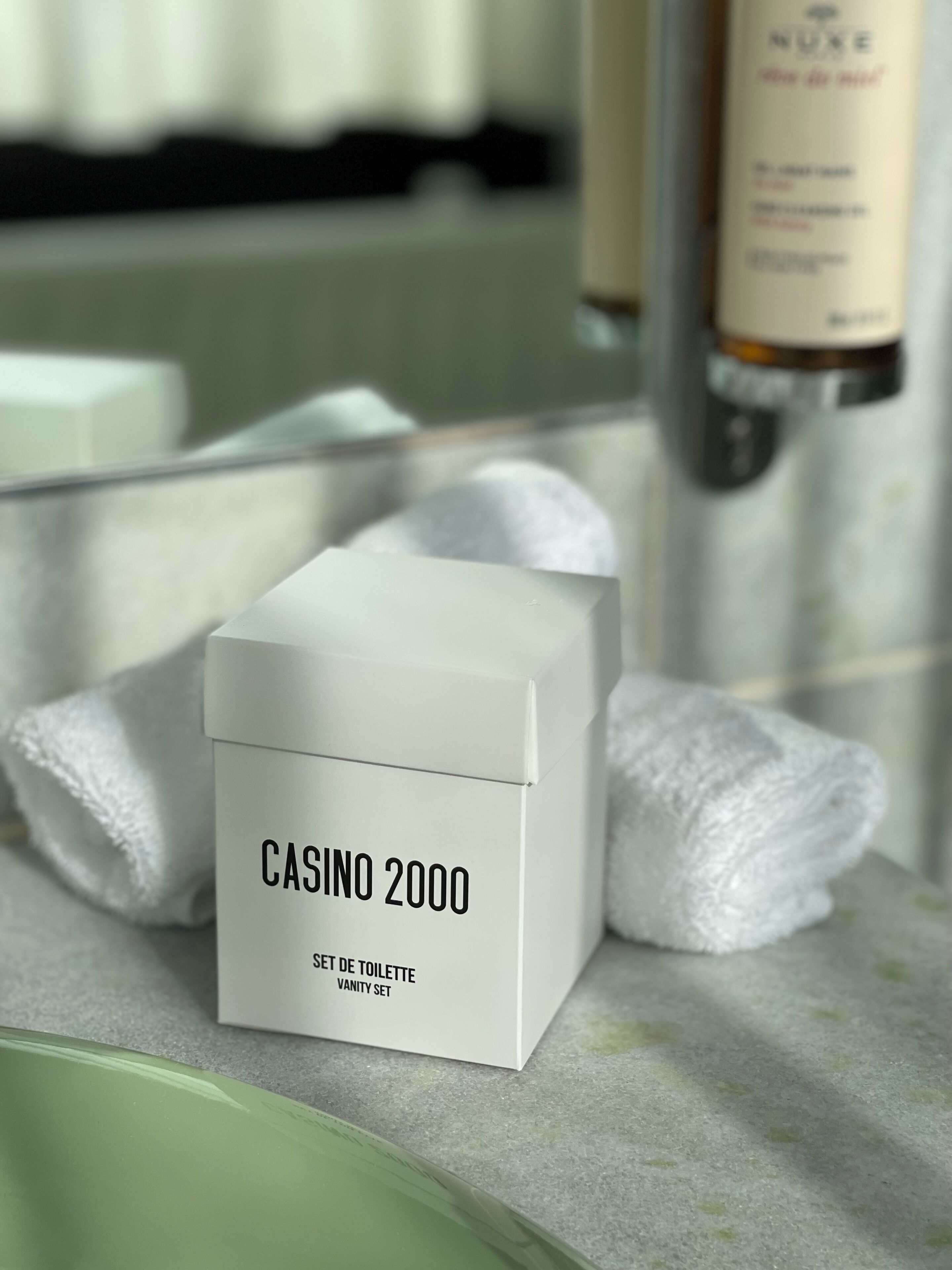 Ein weißes Schminktisch-Set mit dem Aufdruck "CASINO 2000" auf einer Badezimmerablage, im Hintergrund ein Spiegel und eine Flasche.