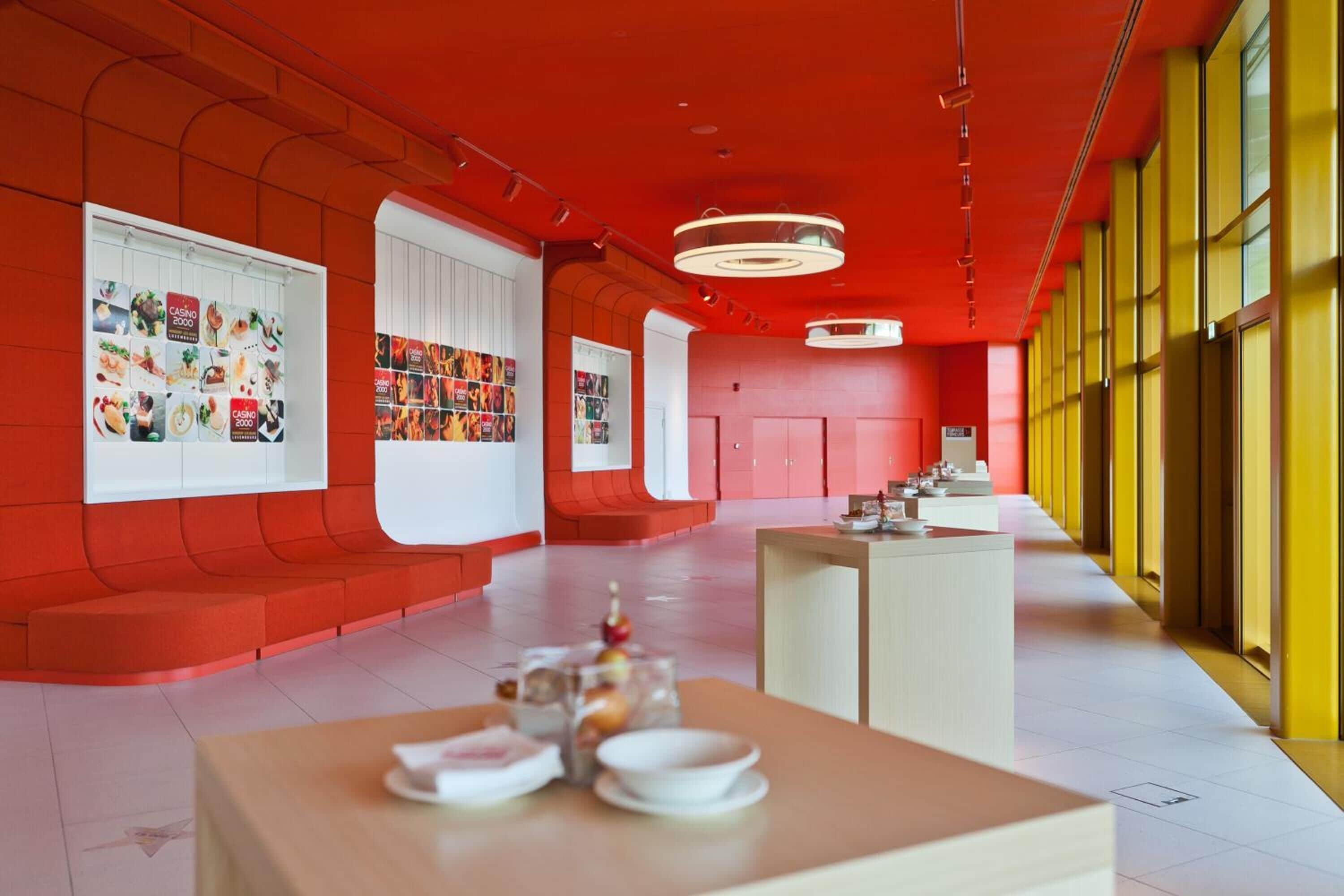 Ein modernes Interieur mit roten und gelben Wänden, einer zentralen Sitzecke und einer langen Theke mit einer Auslage.