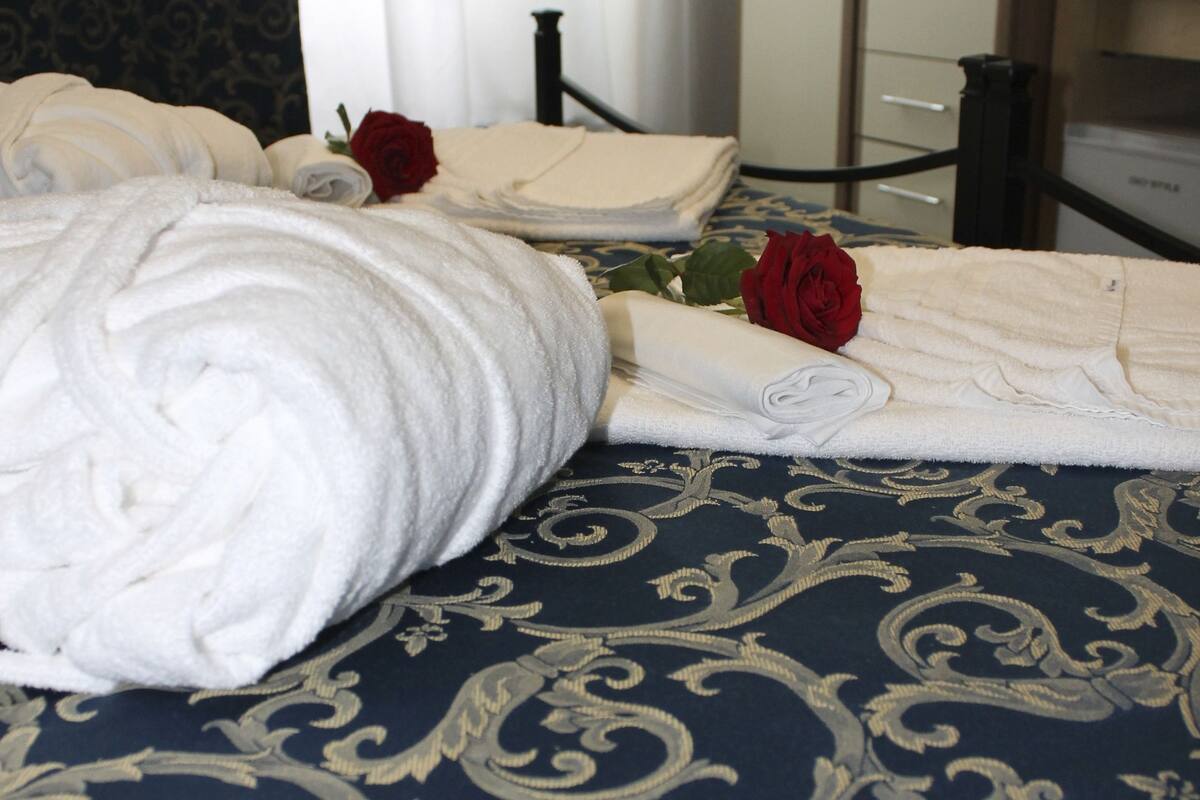 Una cama bien hecha con una toalla blanca enrollada y una rosa roja sobre la almohada.