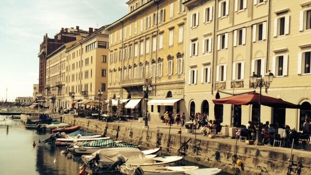 A waterfront with boats docked along the quay, buildings with balconies, and outdoor seating areas. - Hotel Centrale Trieste (Trieste)