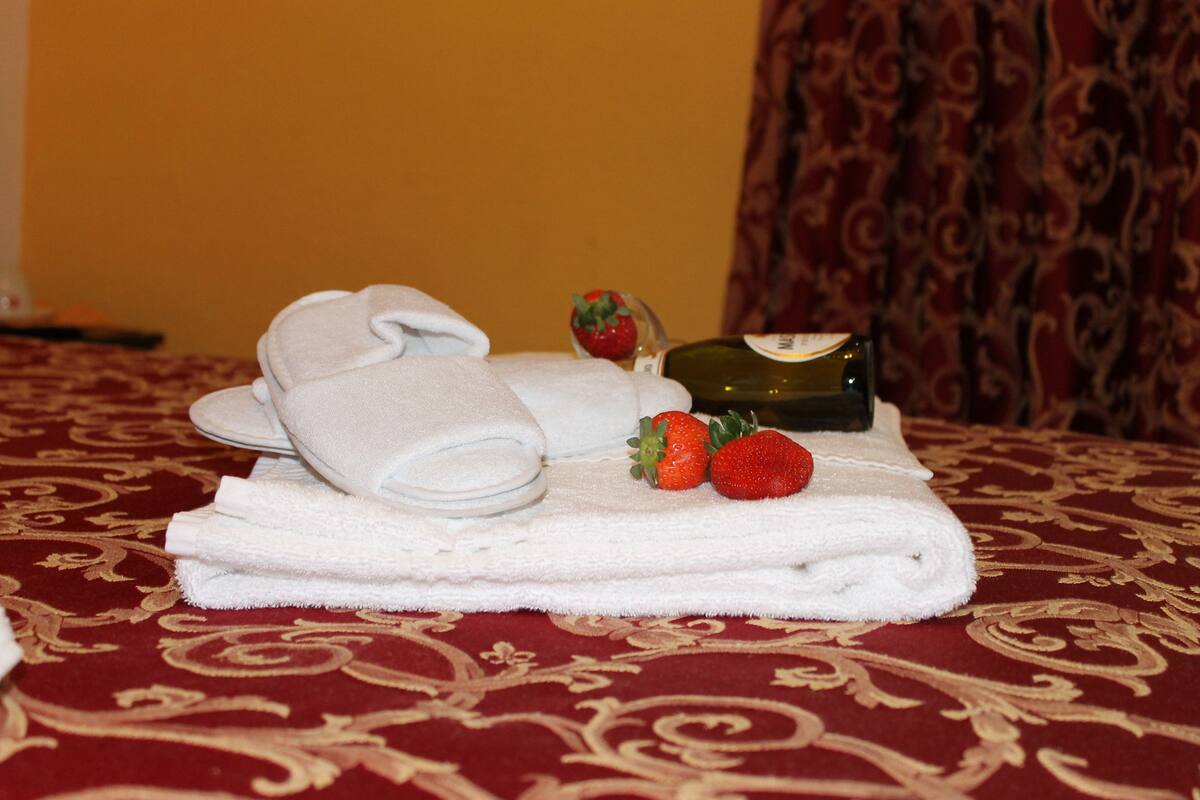 Una toalla blanca doblada con cuidado, una botella de aceite de oliva y dos fresas sobre una cama con una colcha roja estampada.