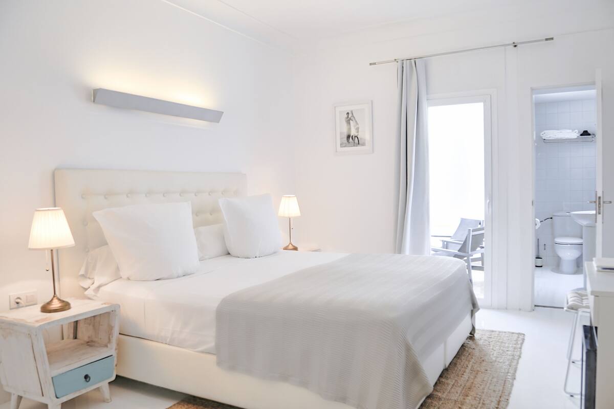 A white bedroom with a large bed, two bedside tables with lamps, a small drawer, a picture on the wall, and an open door leading to a bathroom.