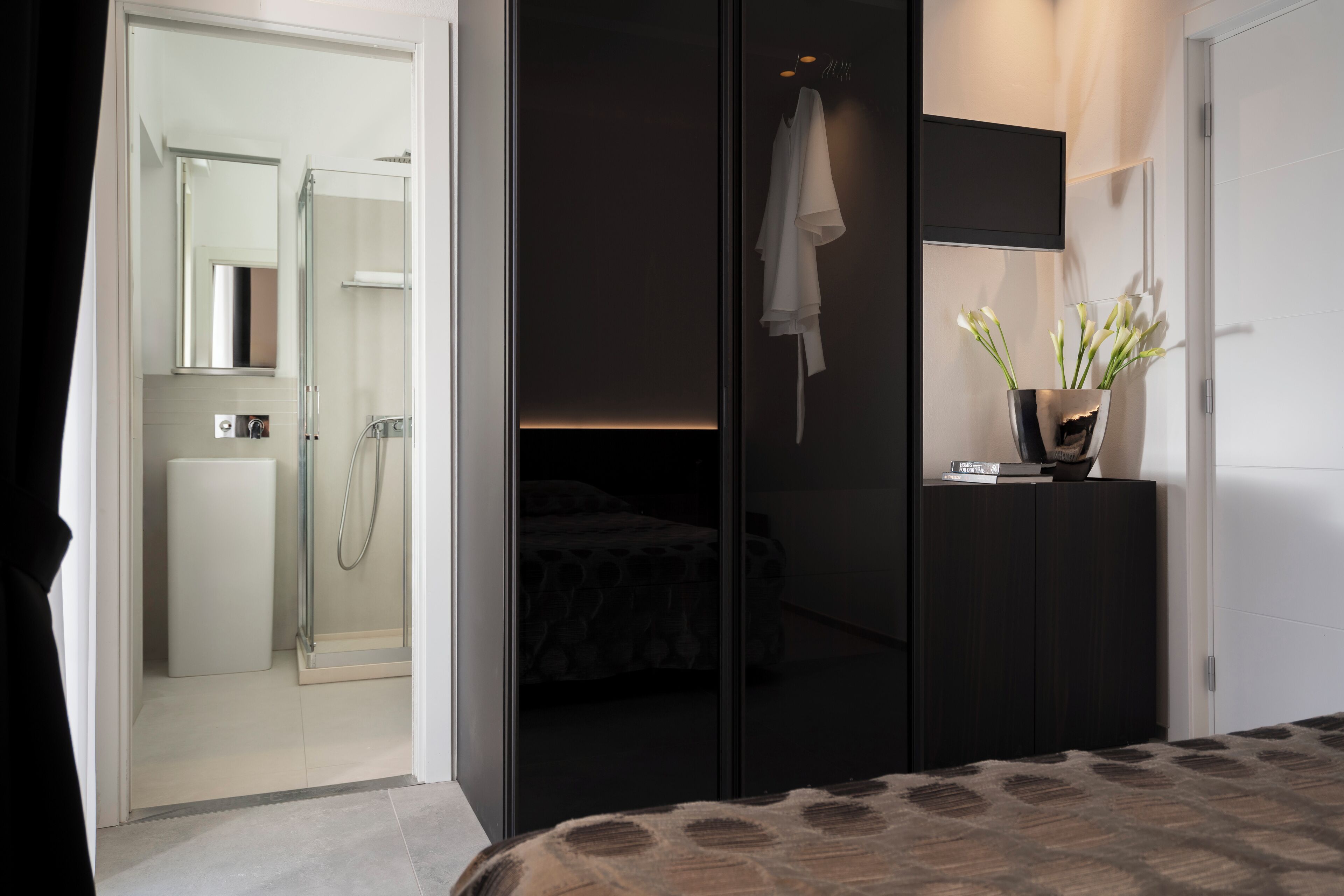 Un dormitorio moderno con cabina de ducha de vidrio, un armario oscuro y una mesita de noche con un florero.