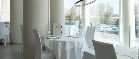 A modern dining area with white tables and chairs, large windows, and a view of parked cars outside.