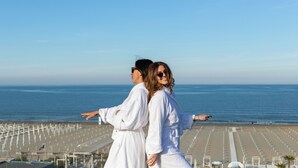 Un couple, vêtu de longues robes blanches, se tient sur un balcon surplombant une plage où s’alignent de nombreux transats.