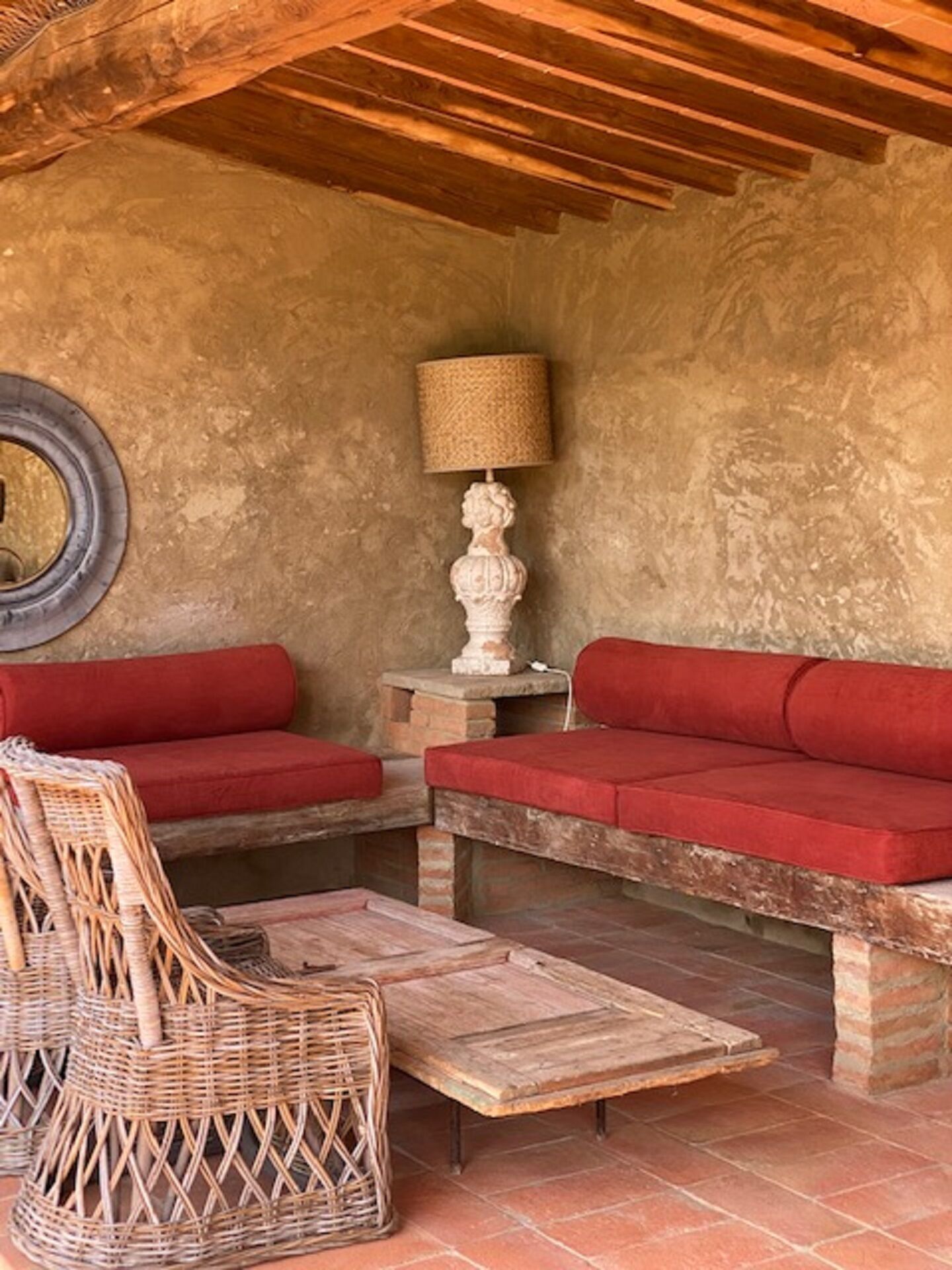 A cozy outdoor seating area with wicker chairs and red cushioned sofas, a woven lamp, and a rustic stone table.