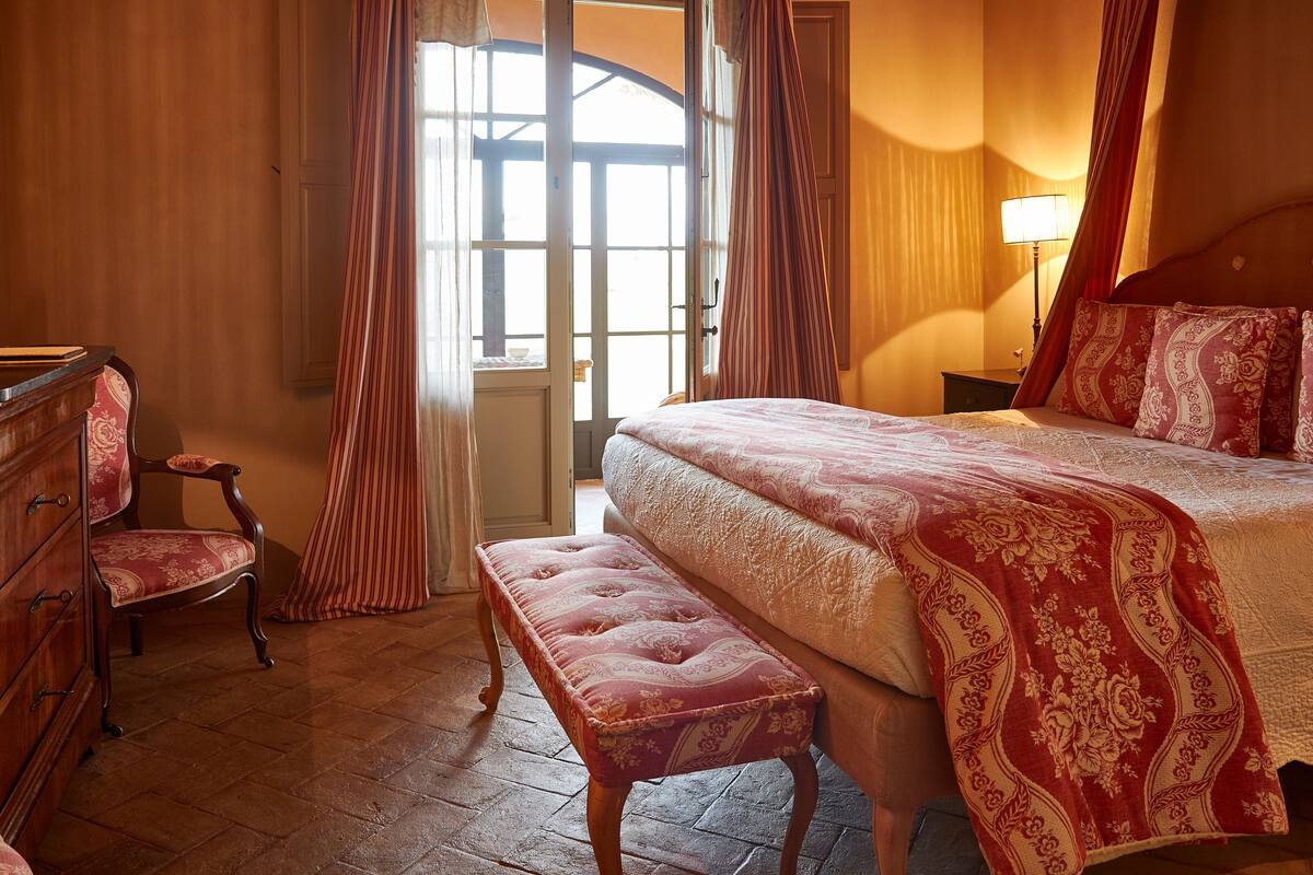 A bedroom with a bed, a bench, a chair, a dresser, and a window with curtains.
