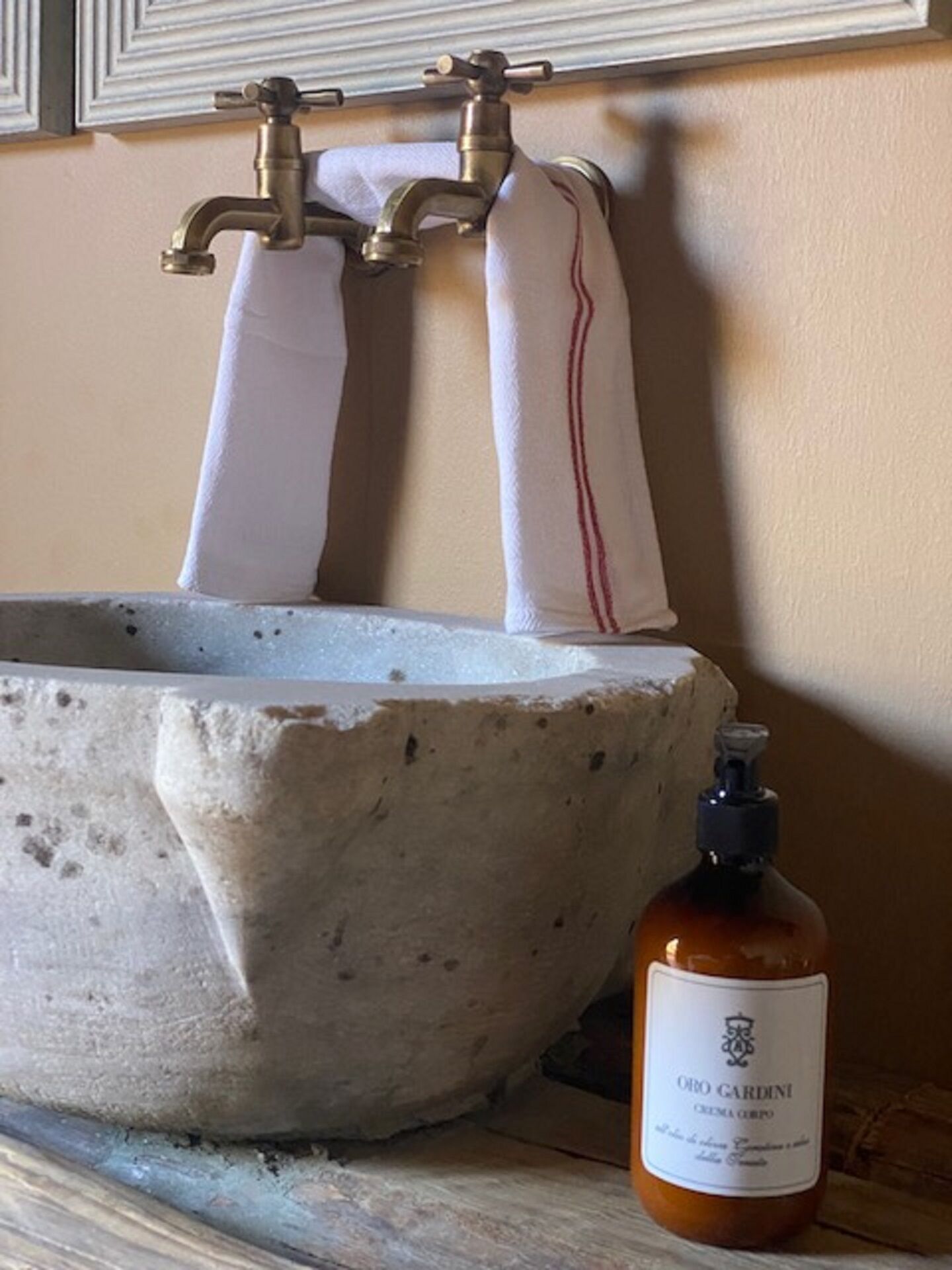Un lavamanos de piedra con dos grifos y una botella de jabón crema ORO GARDINI.