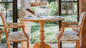A person serving tea at a table with two chairs, a large window, and a view of trees. - Relais Sant'Elena (Bibbona)
