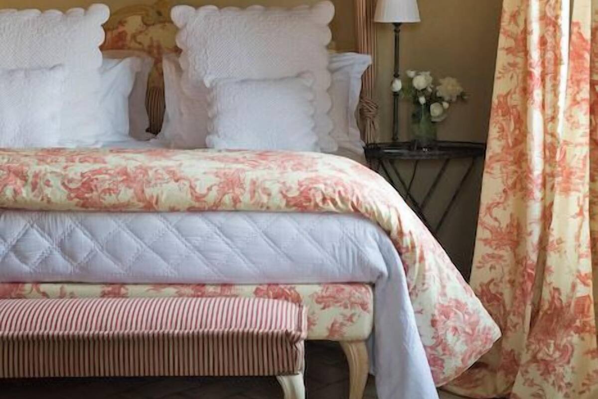 A bed with a floral bedspread and a bench, a nightstand with a lamp, and a floral curtain.