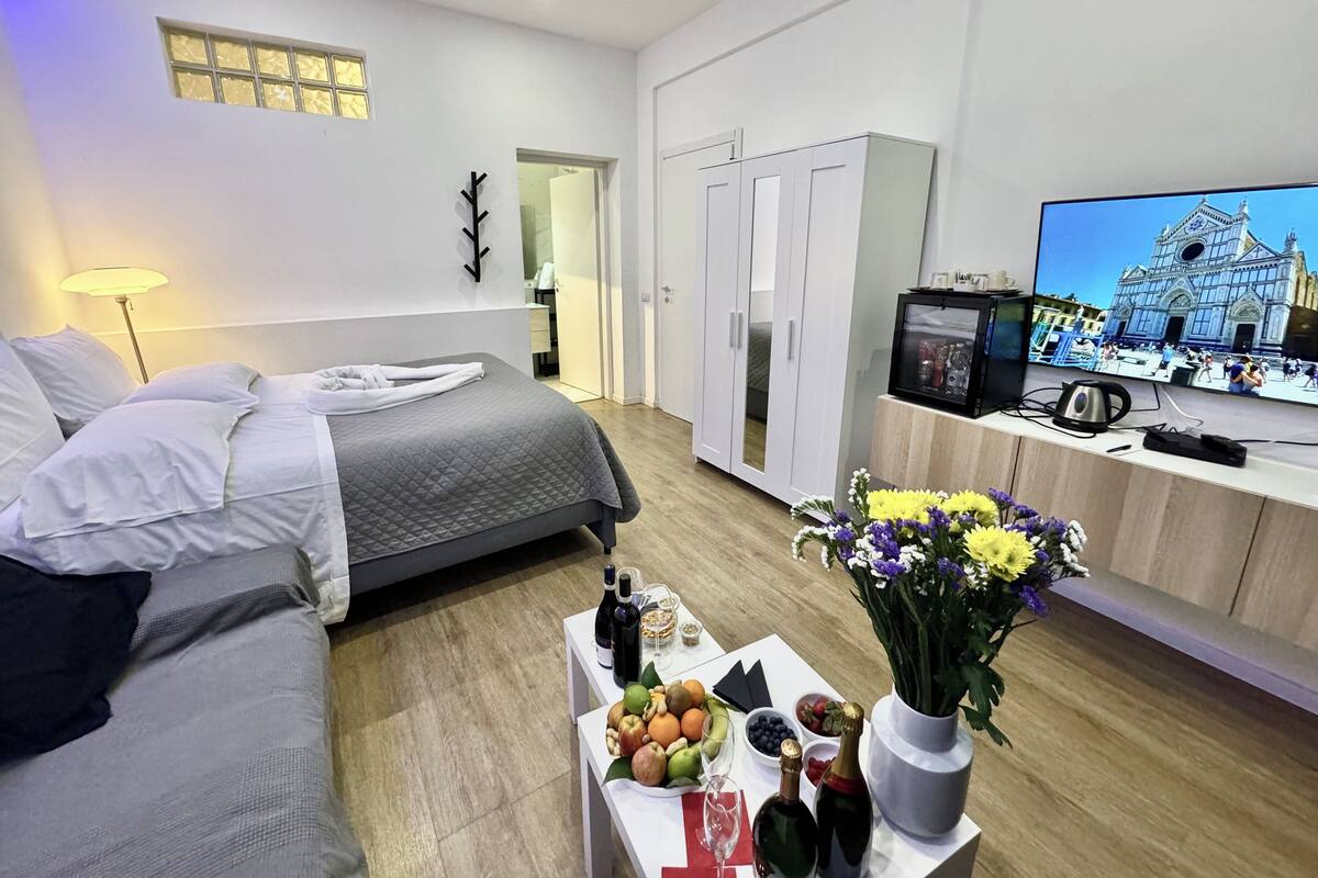 Quarto de hotel moderno com cama, televisão exibindo uma vista urbana, mesa de jantar com frutas e bebidas, e um vaso de flores.