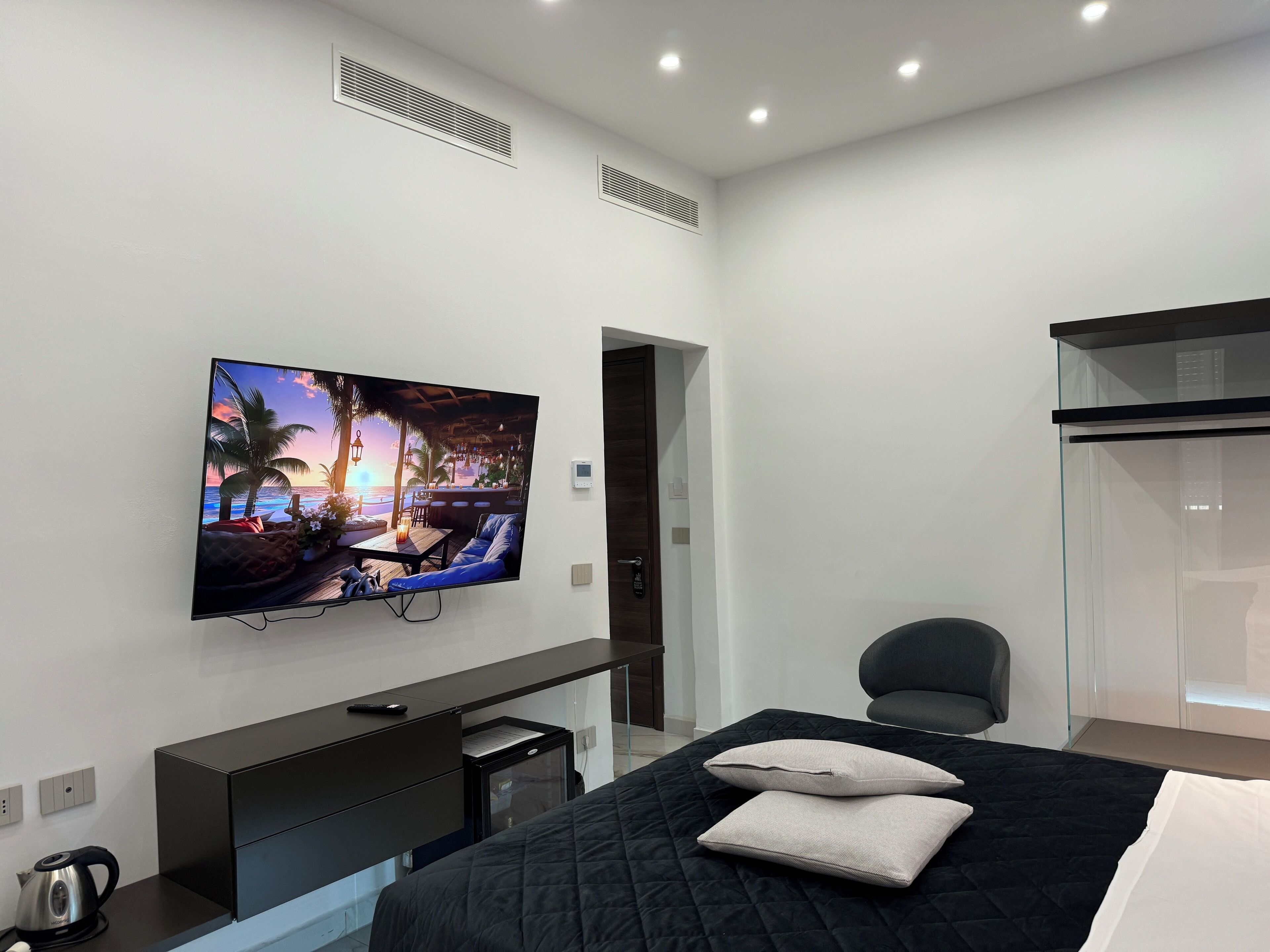 A modern hotel room with a large flat-screen TV mounted on the wall, a bed with a black quilted cover, a desk with a computer, and a chair.