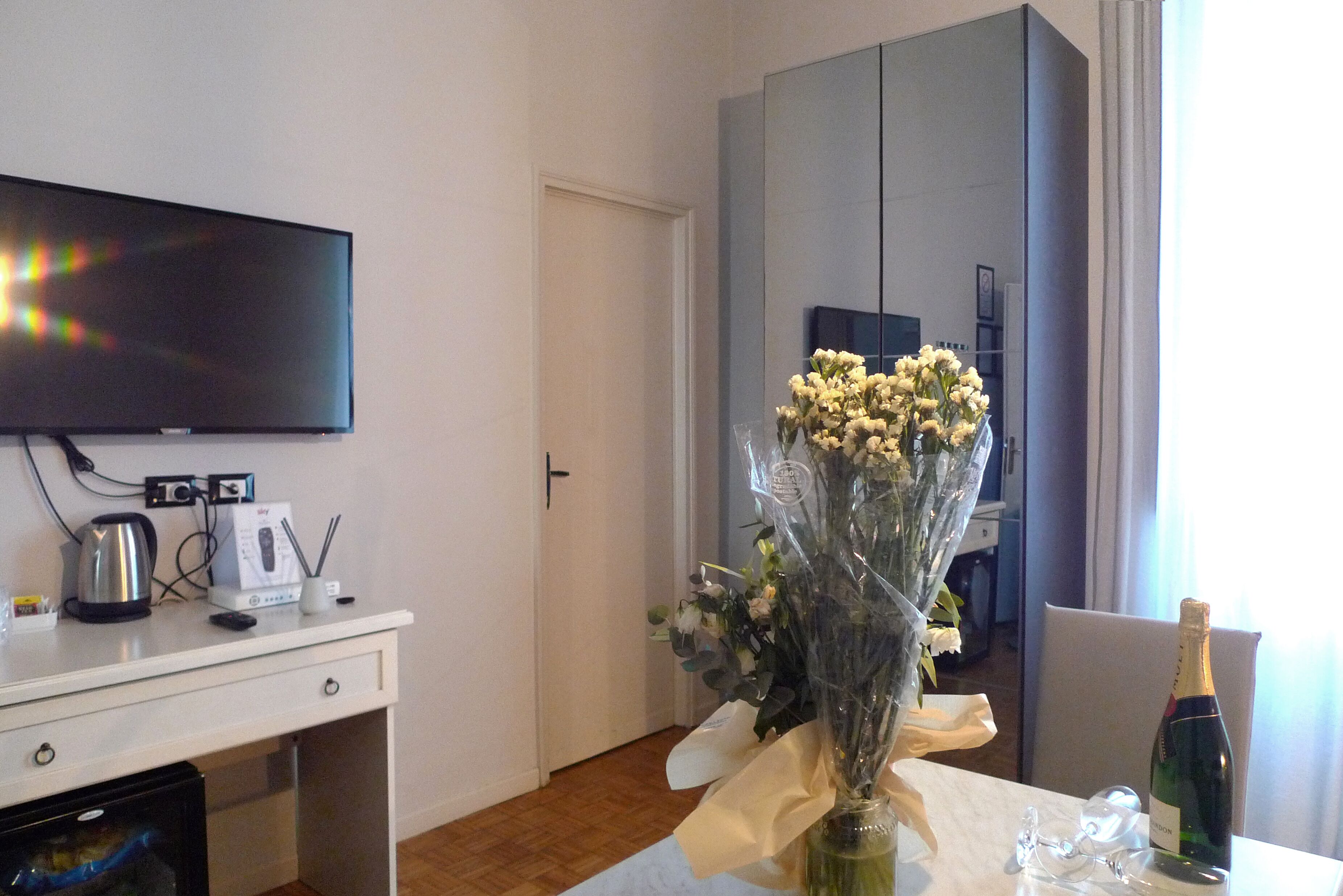 A hotel room with a flat-screen TV, a minibar, a dining table with a vase of flowers, and a bottle of champagne.