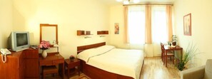 Hotellihuone, jossa on sänky, kirjoituspöytä puhelimineen, televisio ja pieni pöytä, jonka päällä on maljakko täynnä kukkia.