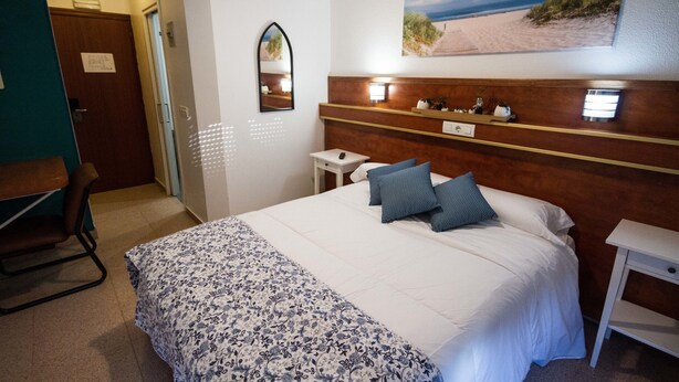 Een hotelkamer met een bed, een bureau, een stoel, een nachtkastje, een spiegel en een wanddecoratie met een strandthema.