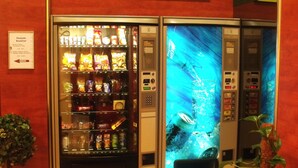 Een automaat met diverse snacks en drankjes, een digitaal scherm en een bord met de openingstijden voor ontbijt.