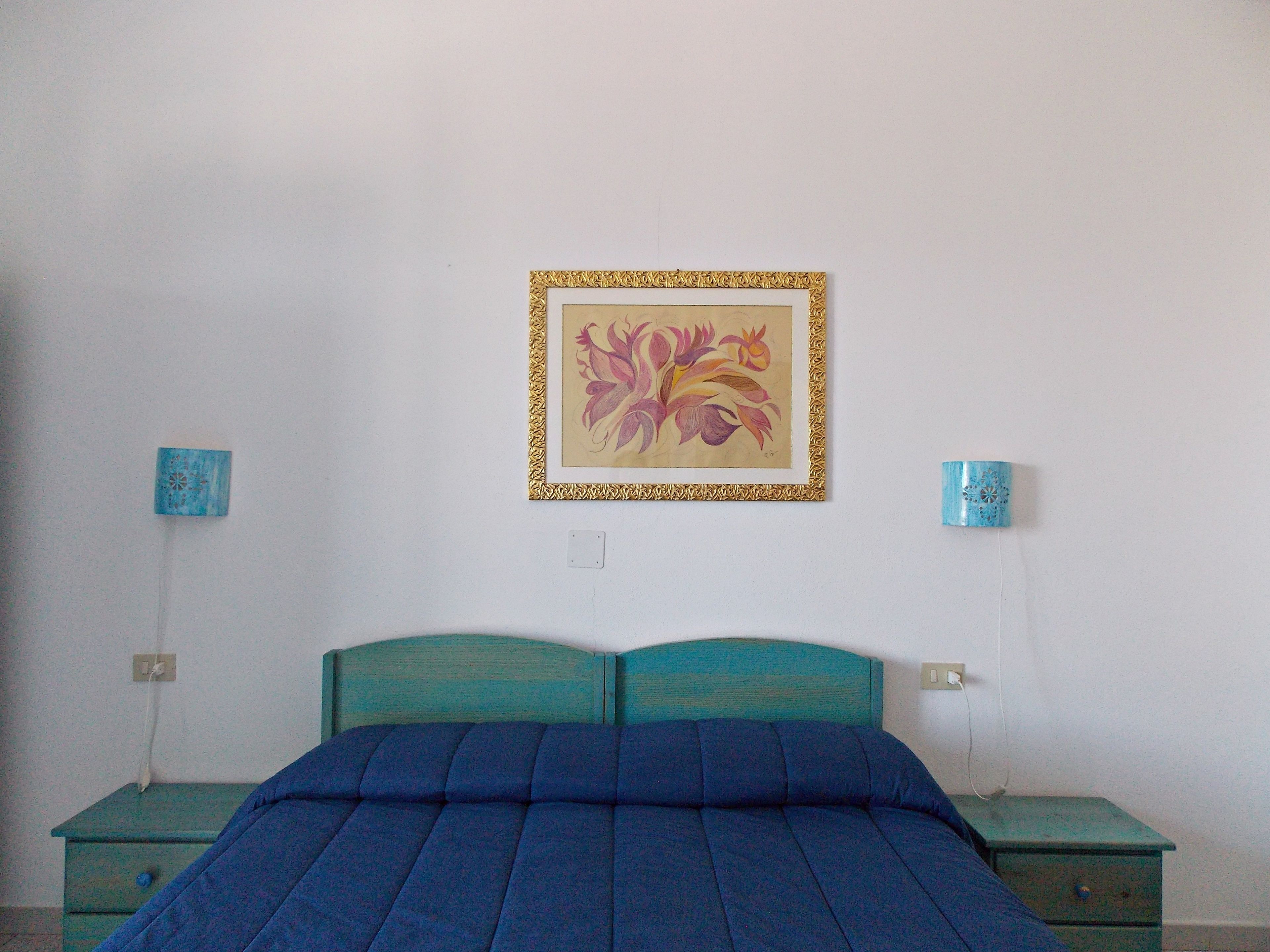 A bedroom with a teal headboard, a blue bedspread, and a framed floral artwork on the wall. There are two bedside tables with blue lamps.