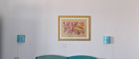 A bedroom with a teal headboard, a blue bedspread, and a framed floral artwork on the wall. There are two bedside tables with blue lamps.