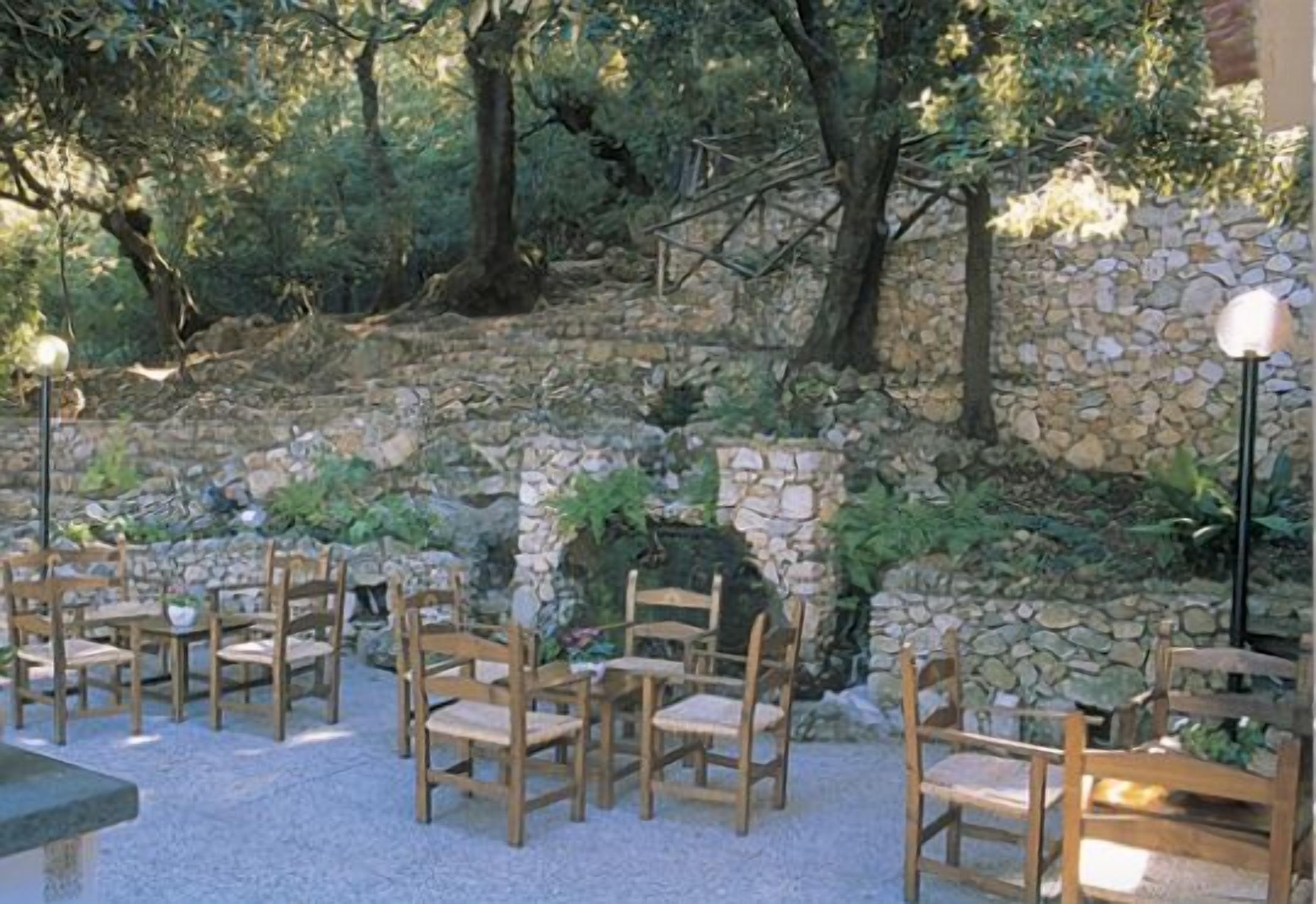An outdoor seating area with wooden chairs and tables, surrounded by stone walls and greenery.