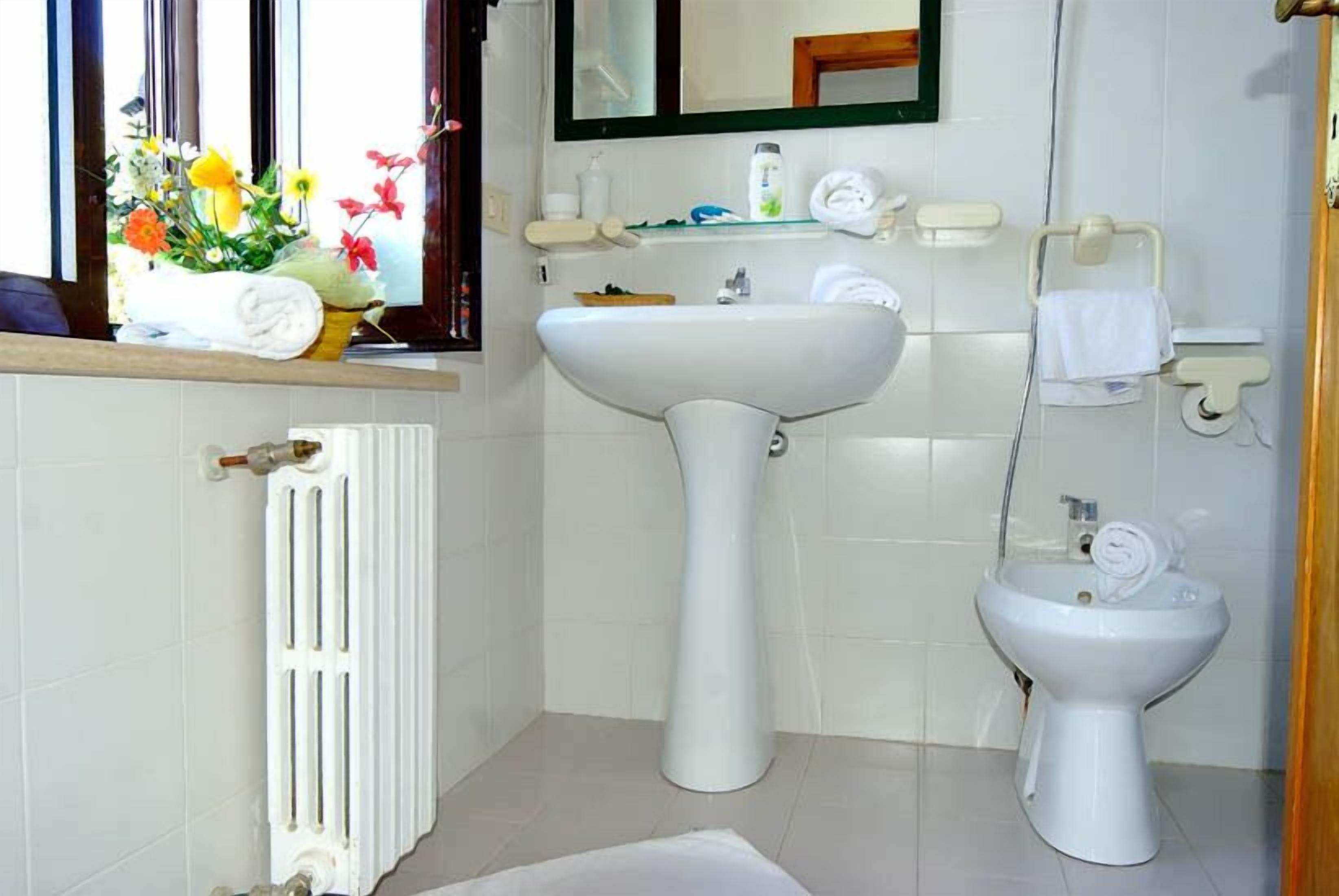 Une salle de bain avec un lavabo sur colonne, un bidet, un miroir et une fenêtre agrémentée d’un arrangement floral.