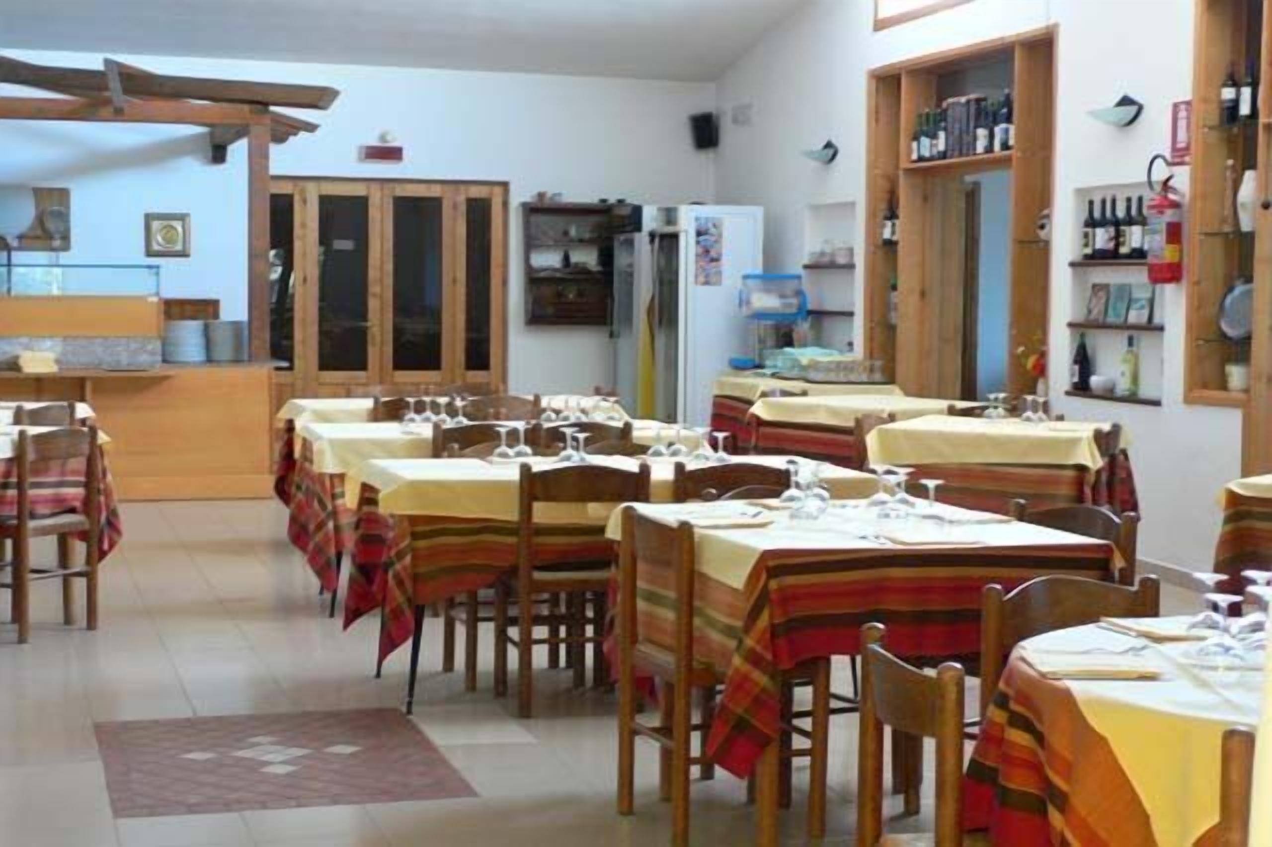 Un comedor con varias mesas y sillas, una zona de cocina con refrigerador y estantes, y armarios de madera.