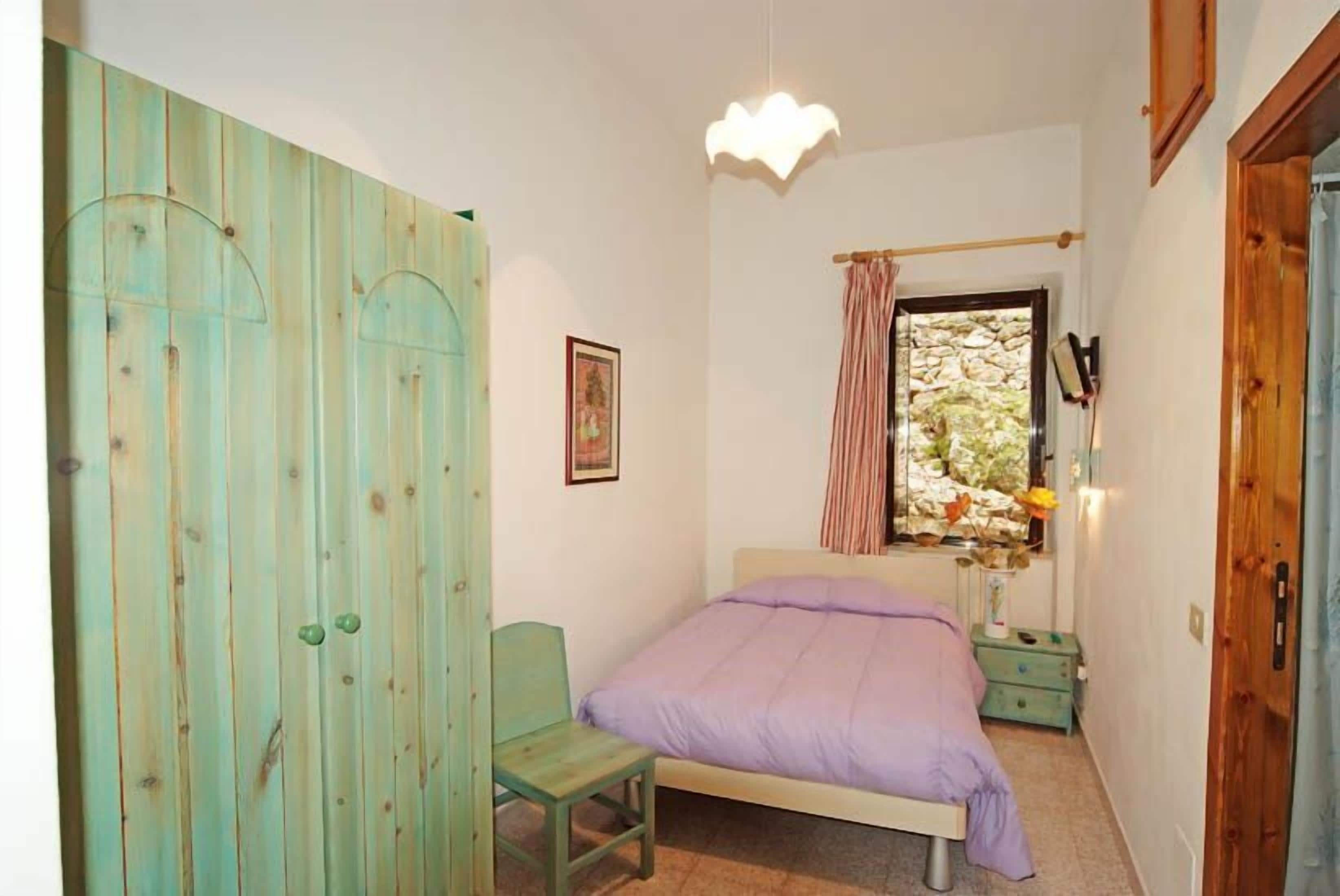 A bedroom with a bed, a chair, a wardrobe, a window with curtains, and a wall-mounted light fixture.