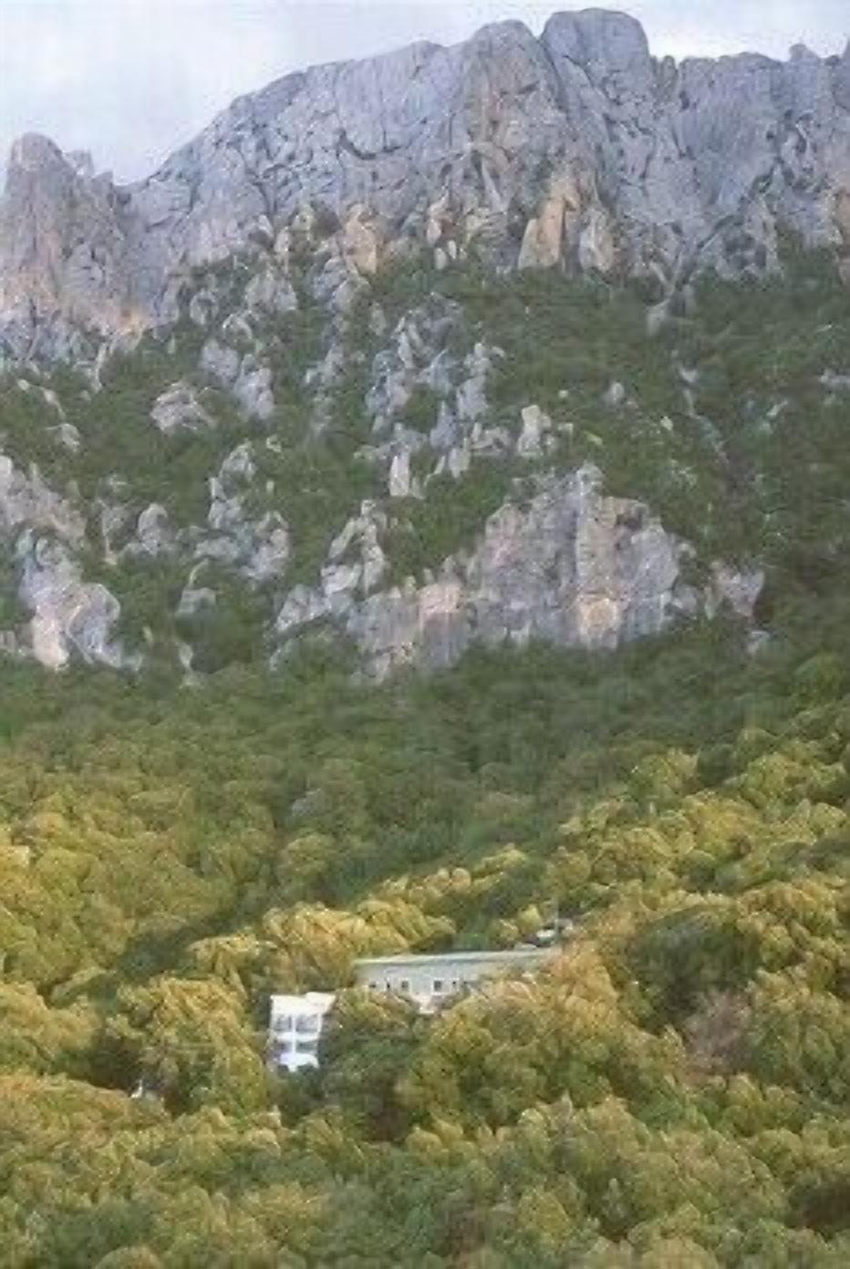 A mountain with rocky terrain and a building surrounded by dense green foliage.