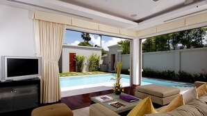 A modern living room with a flat-screen TV, comfortable seating, a coffee table with magazines, and a view of a swimming pool and greenery outside.