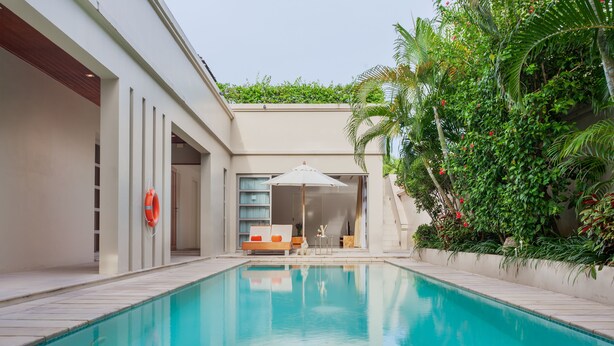 A swimming pool with a sun lounger and a small table, surrounded by greenery and a building with a modern design.