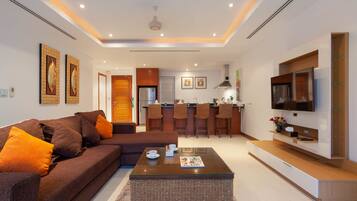 A modern living room with a sofa, coffee table, and a TV. There is a kitchen area with a bar and a refrigerator.